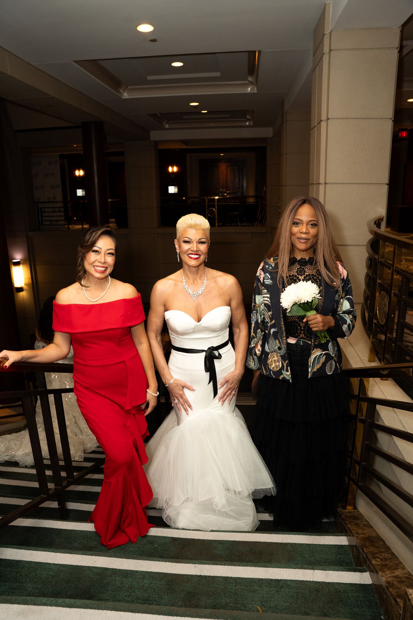 Group photo of models in formal gowns posing together in an elegant venue hallway.