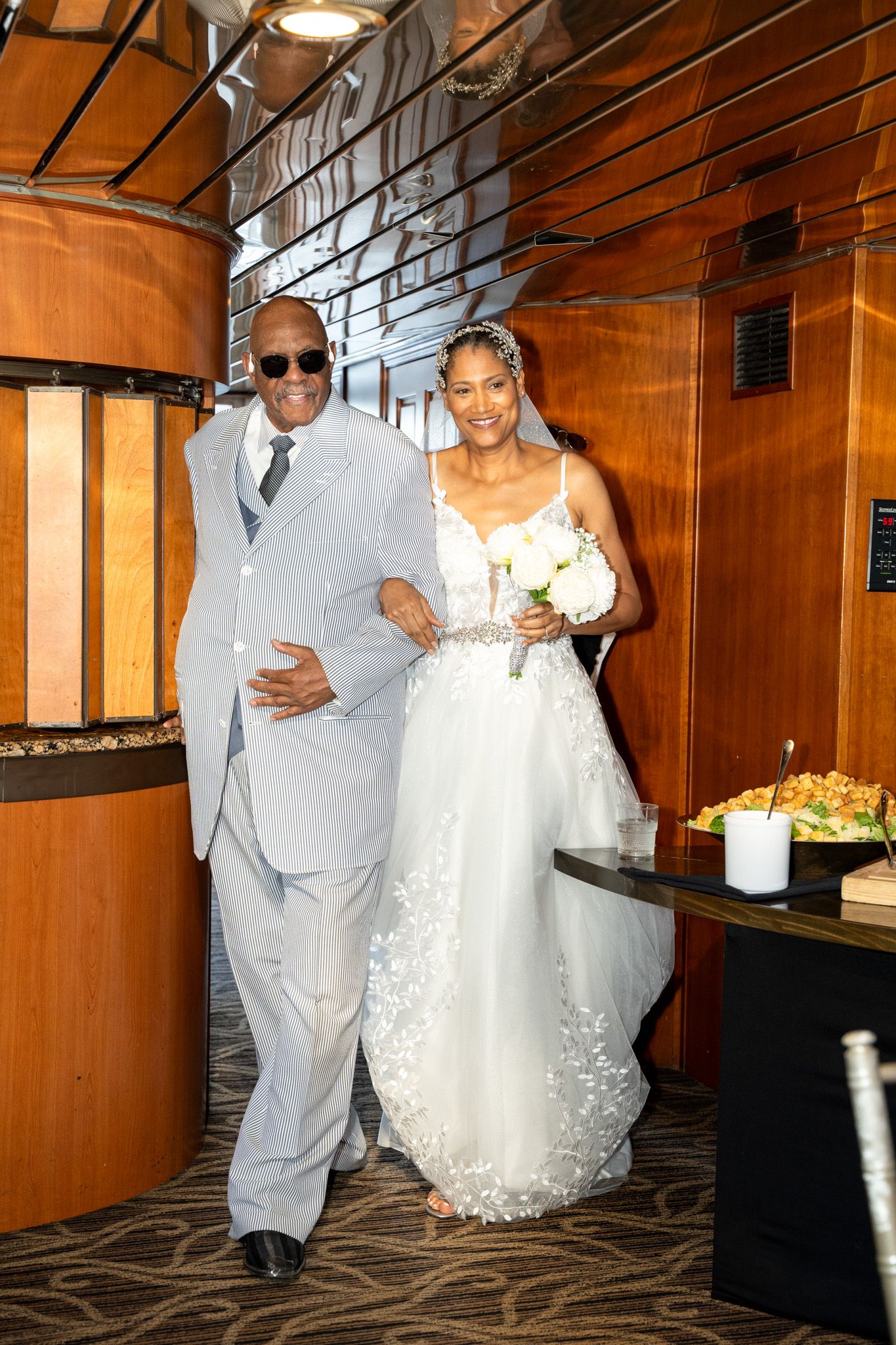 A couple walks down a wood-paneled hallway at a wedding ceremony, with the bride wearing a white lace gown.