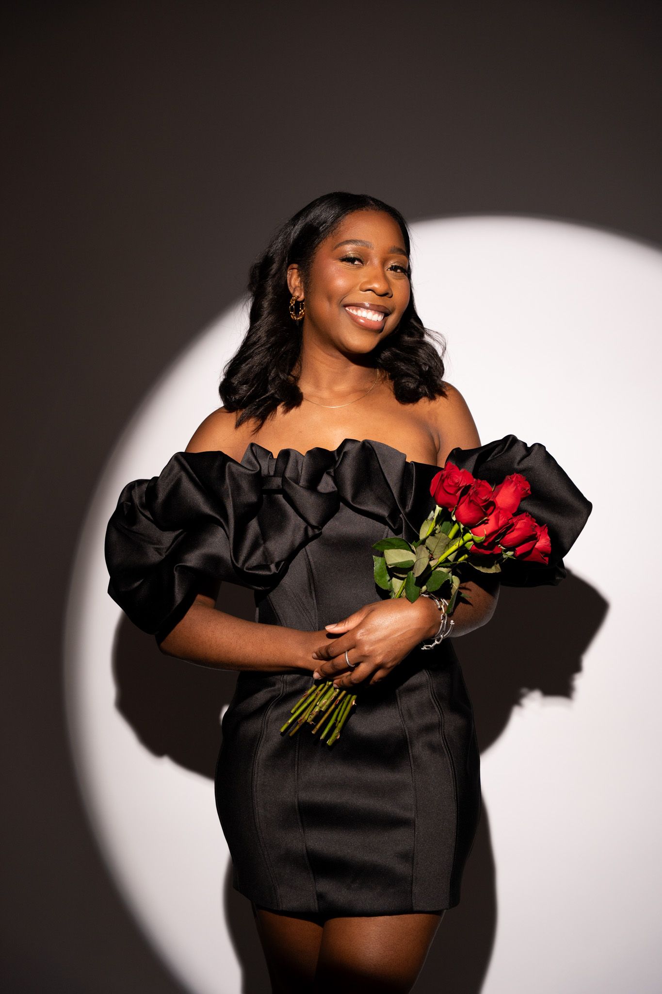 Woman in black off-shoulder mini dress holds red roses against dark background with dramatic spotlight effect.