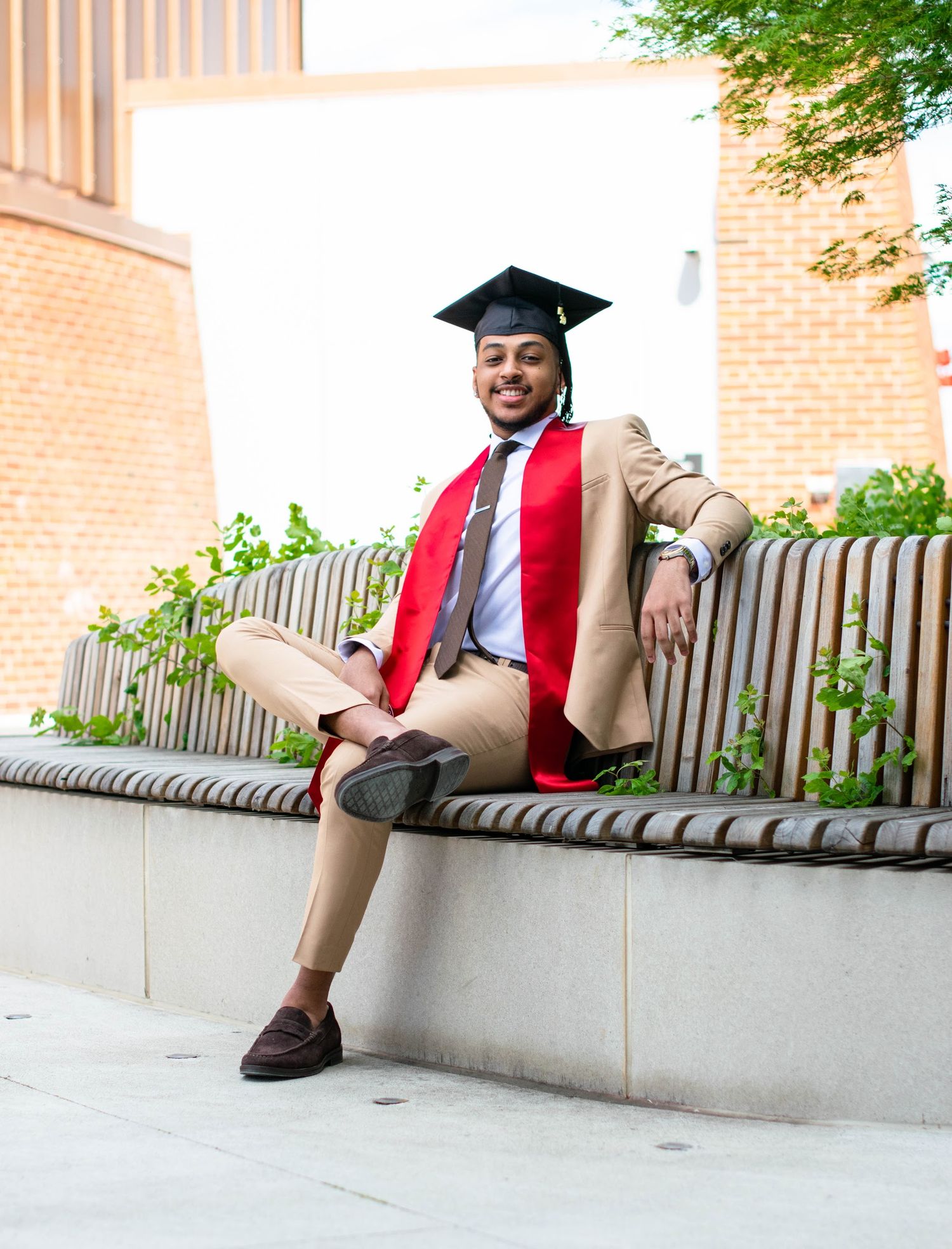Graduate in tan suit and red stole relaxes on outdoor bench during graduation photoshoot.