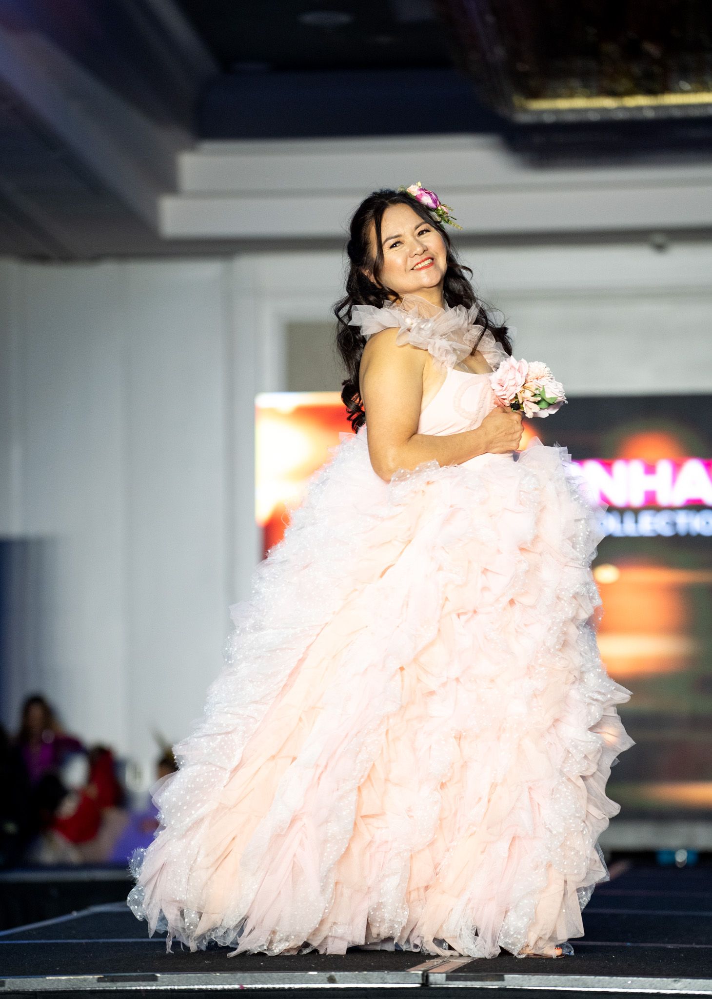 Model in pale pink ruffled gown glides across fashion runway stage.