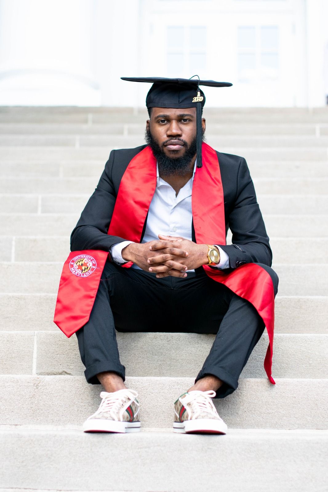 Graduate in black suit and red stole sits contemplatively on concrete steps wearing casual sneakers.