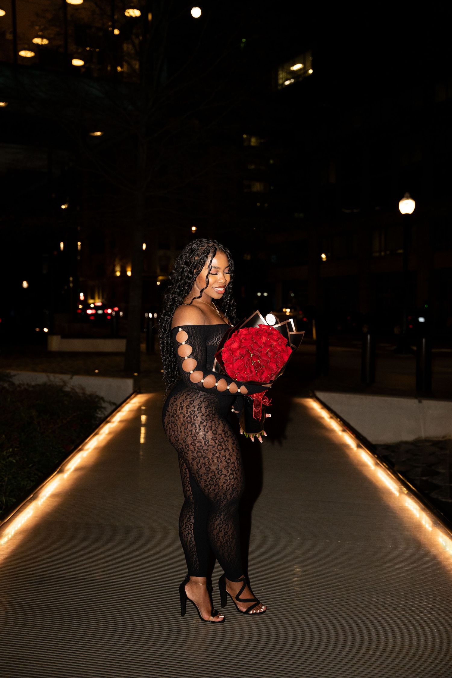 A figure in a black lace dress poses with red roses against an illuminated pathway at night.