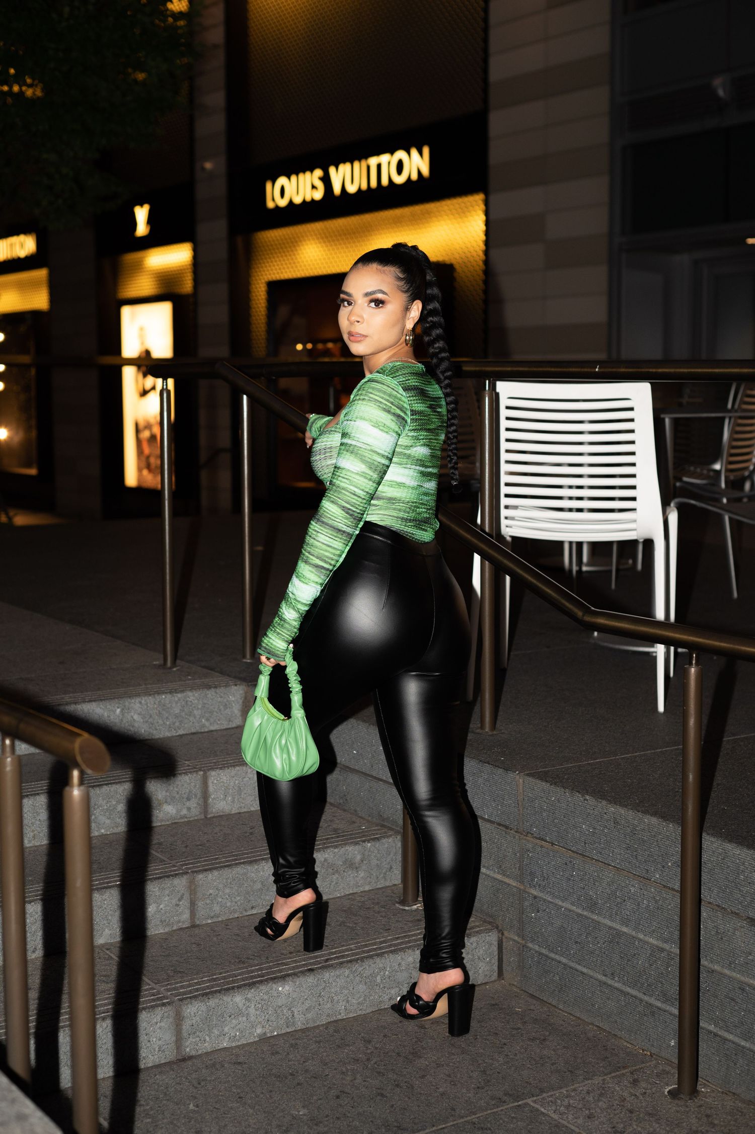 Fashionable woman in metallic green top and leather pants poses on steps outside Louis Vuitton store at night.