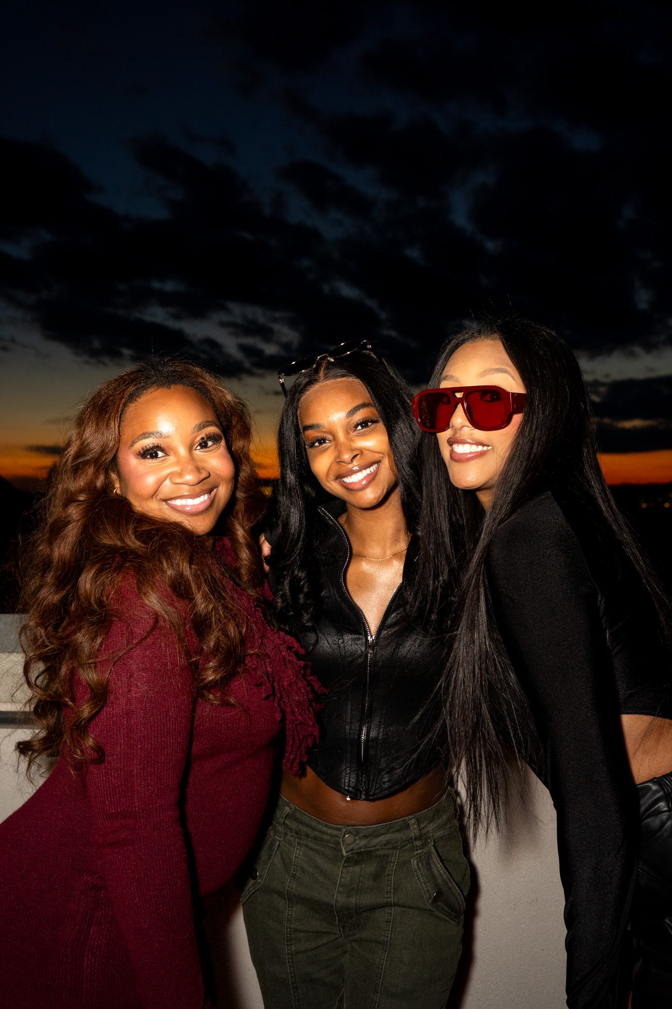 Three friends pose together at sunset wearing stylish outfits.