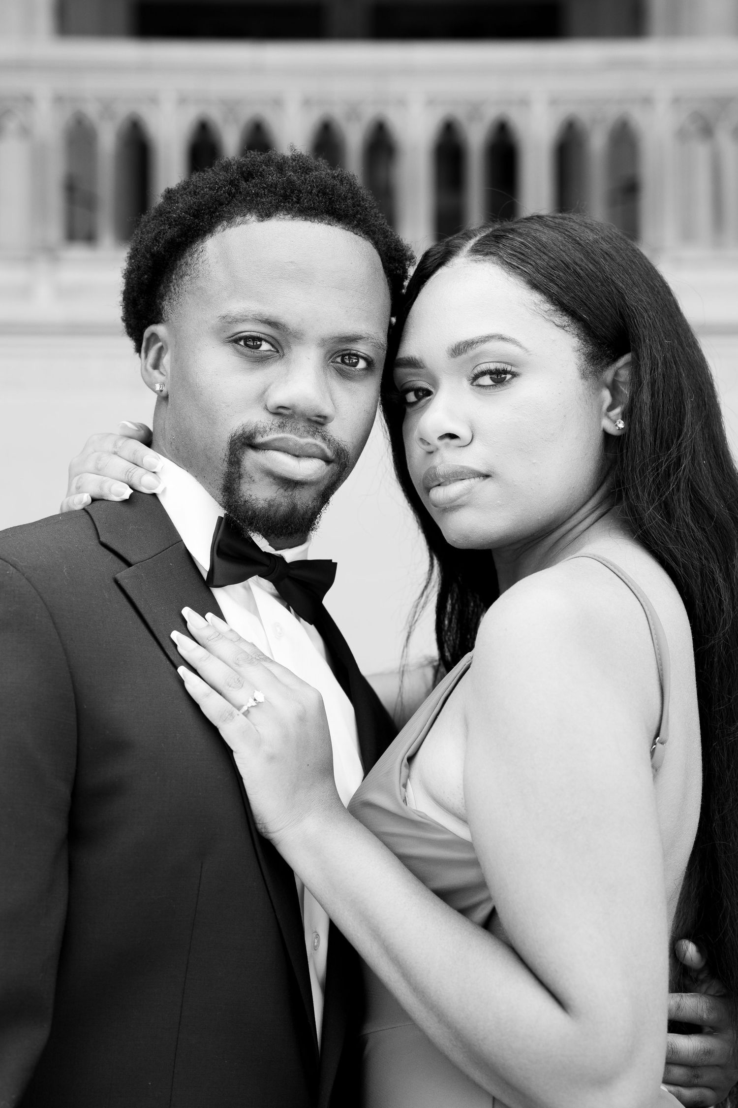 Formal portrait of couple in black tie attire against architectural backdrop.