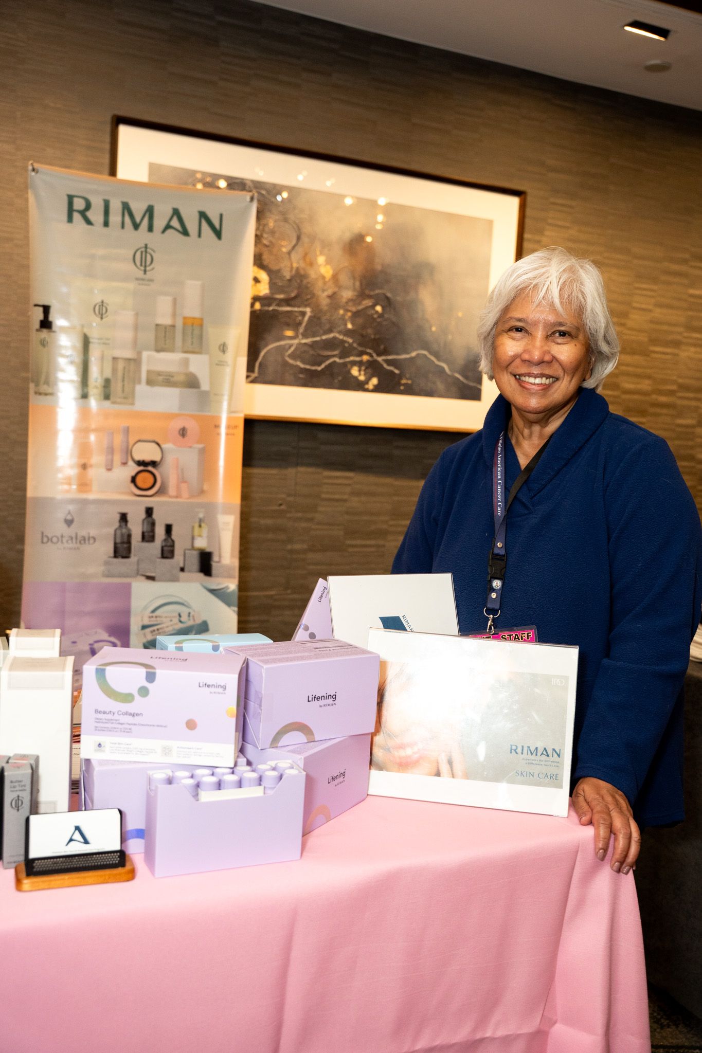 Display table with pink tablecloth featuring Riman beauty products and promotional materials.