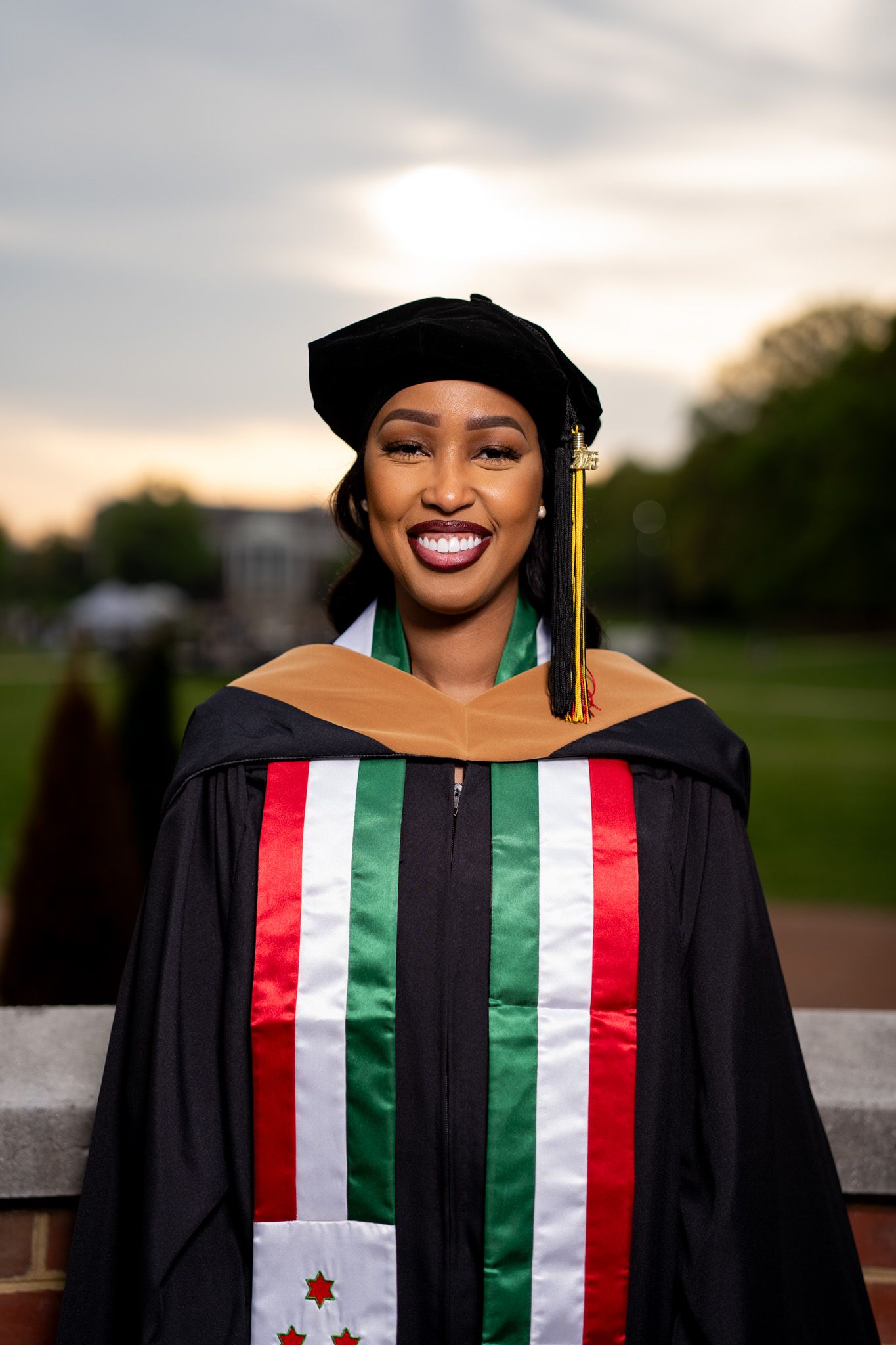 Graduate wearing academic regalia with colorful stoles against evening sky.
