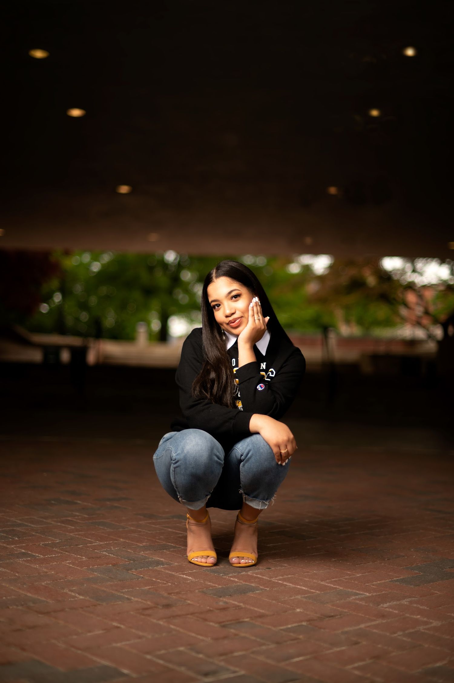 Graduate in dark attire poses on brick pavement in evening lighting.