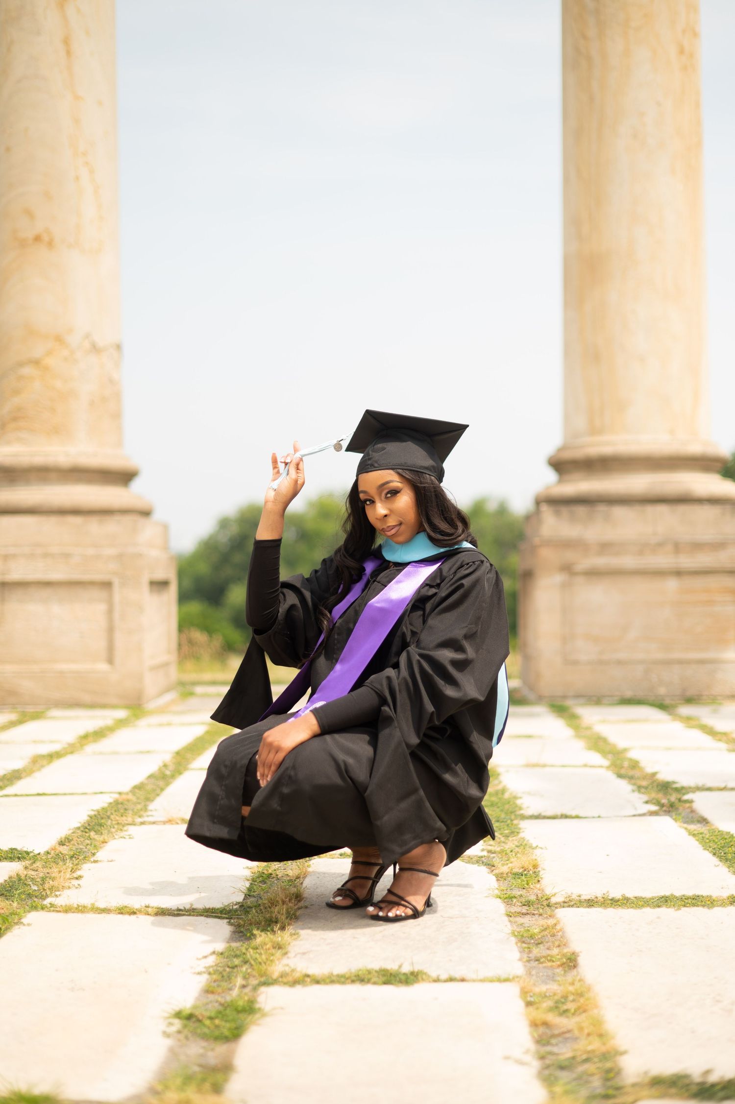 A graduate in cap and gown poses playfully on stone steps between classical columns.