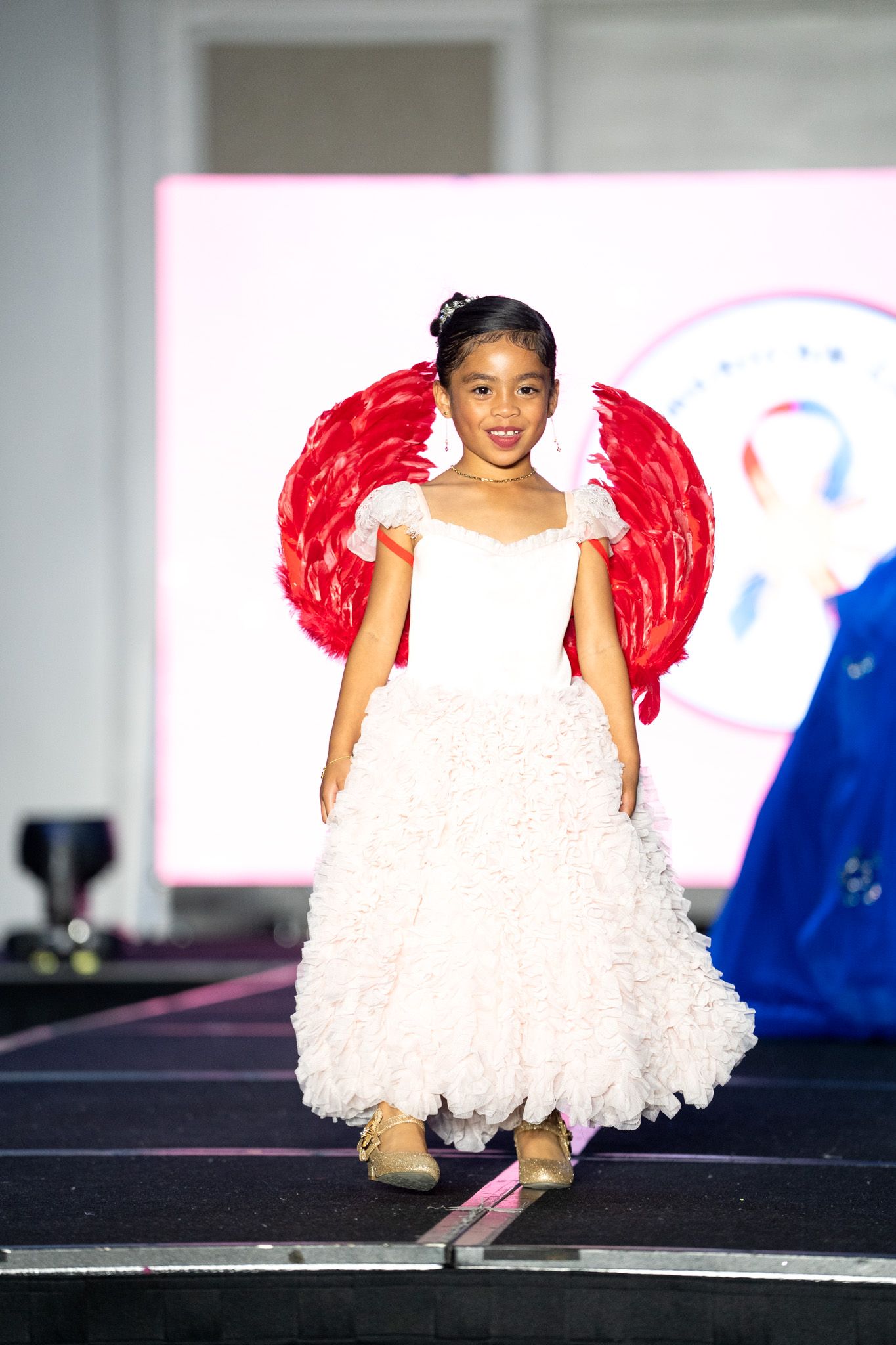 Performer in white ruffled dress with red wings poses confidently on fashion runway.