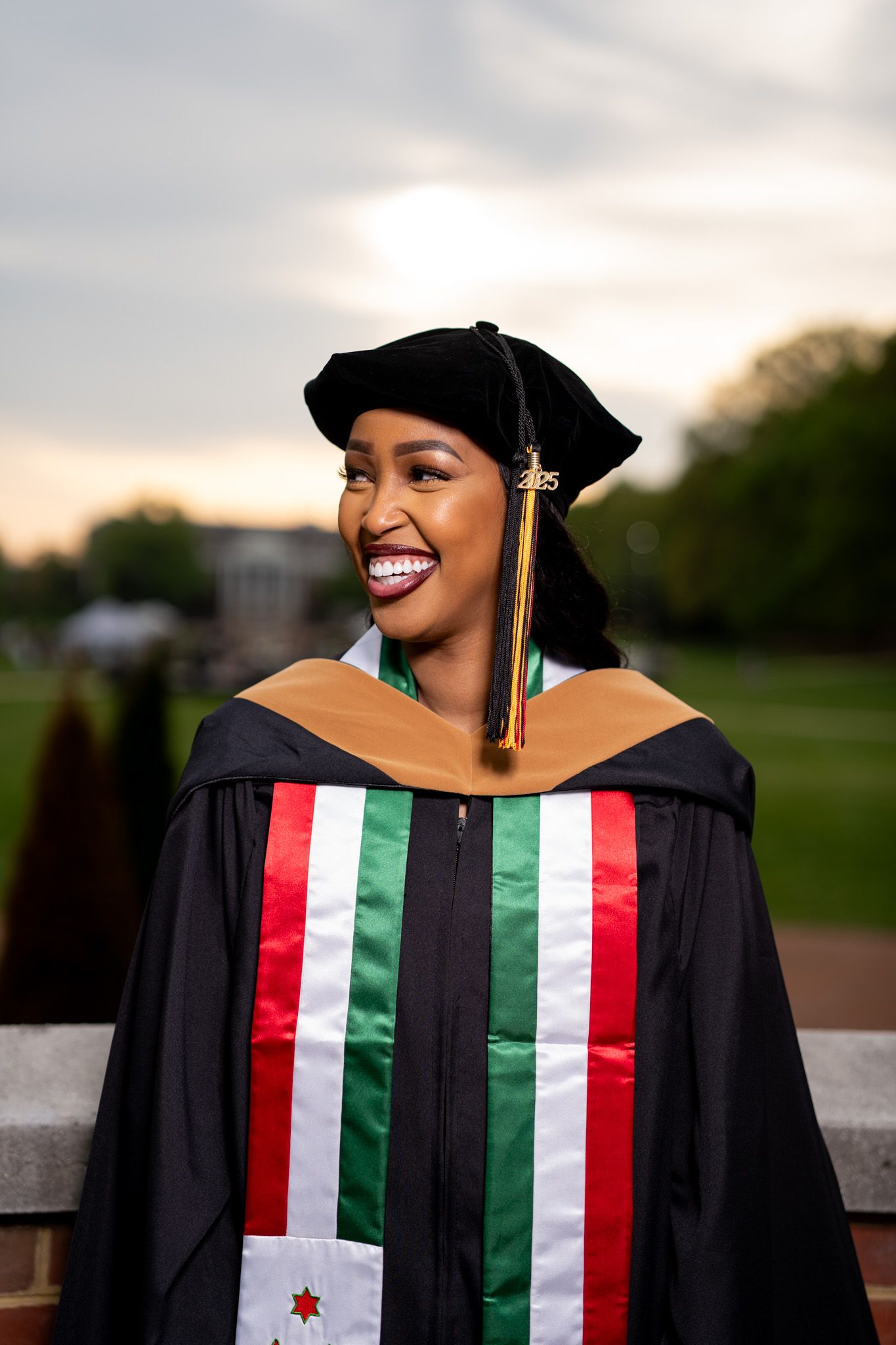 Graduate in decorated academic regalia smiles joyfully during sunset photo session.