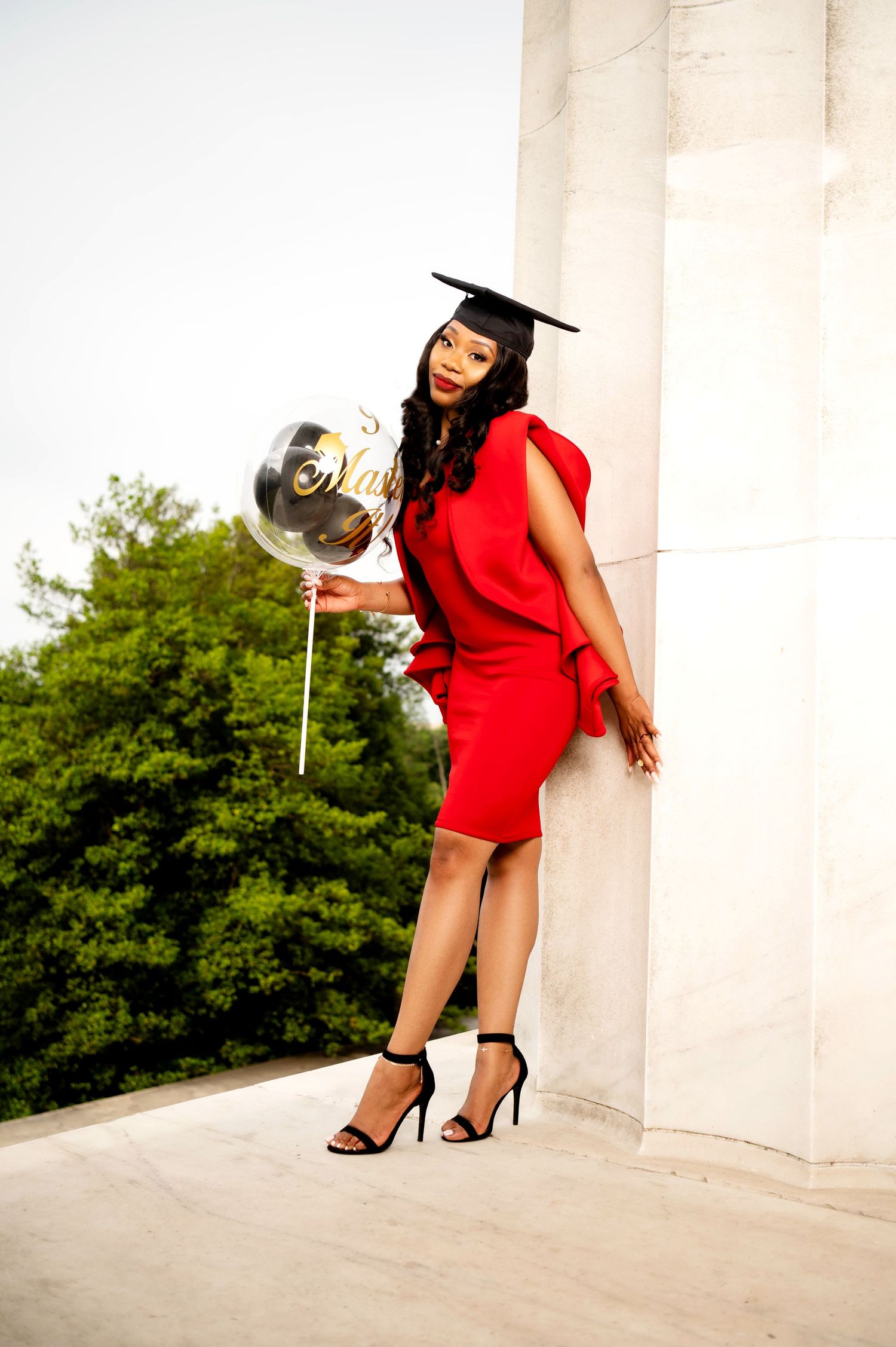 Graduate in red dress poses with graduation balloon by white pillars.