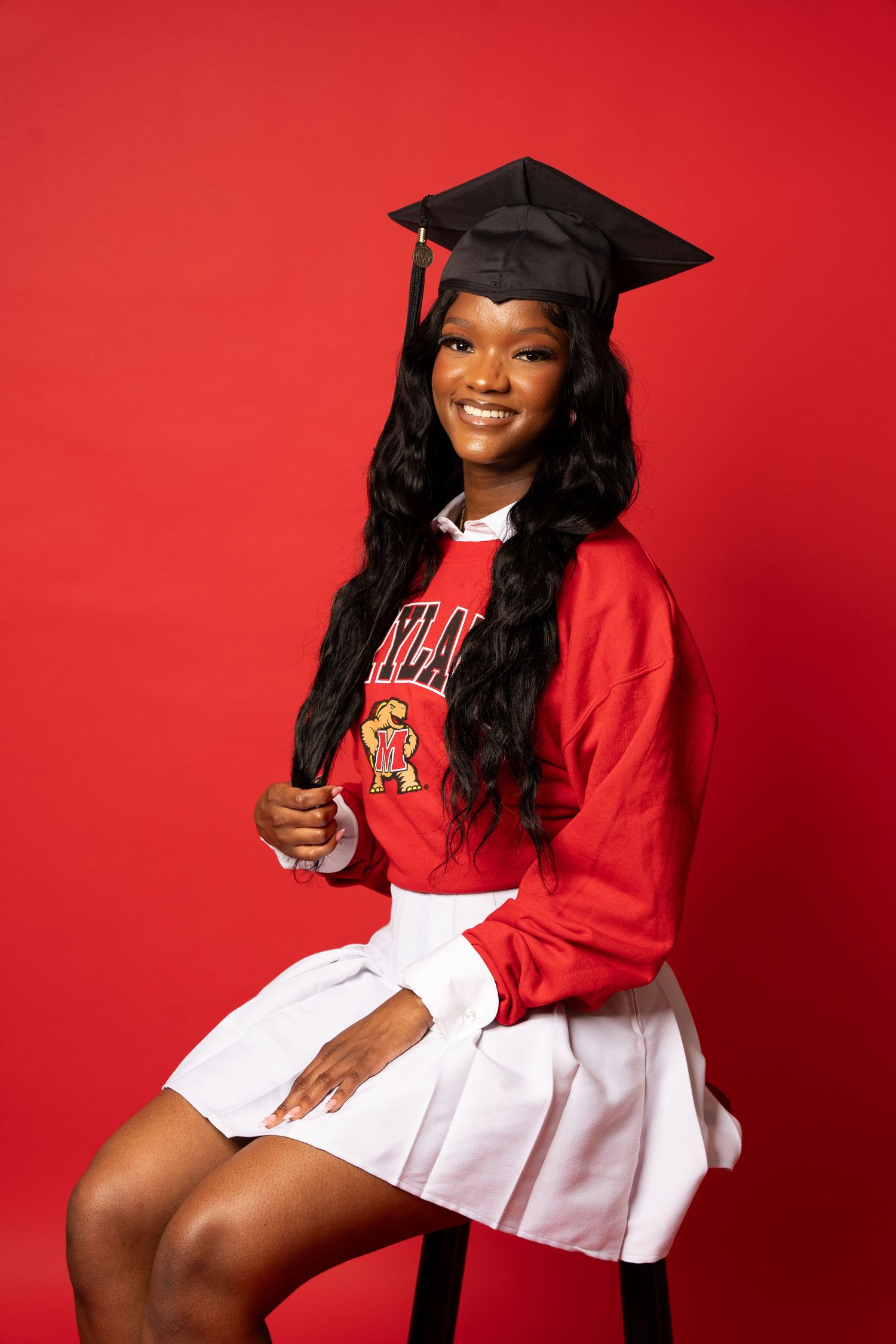 Graduate in red university sweatshirt and white pleated skirt poses against red background.