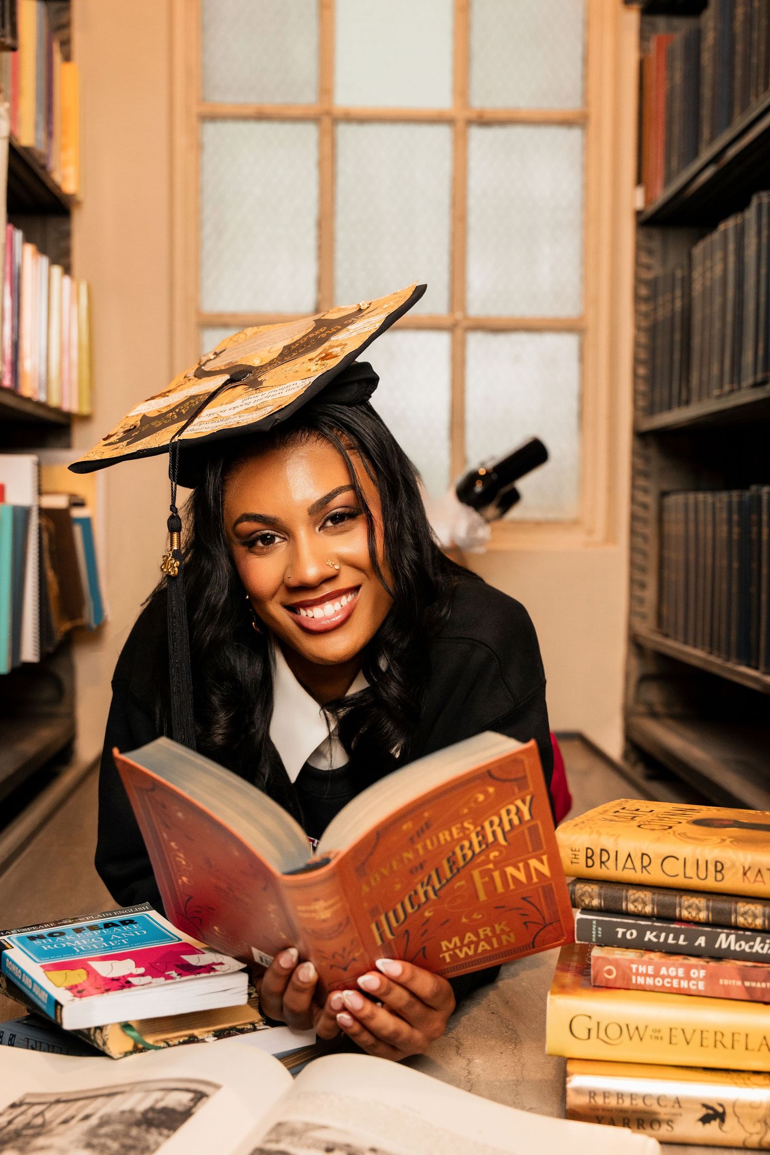 Graduate in cap and gown smiling while reading Huckleberry Finn by Mark Twain in a library.
