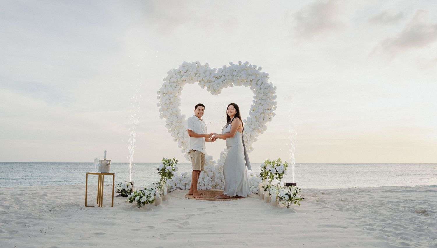 Couple holds hands in front of white flower heart arch on sandy beach.