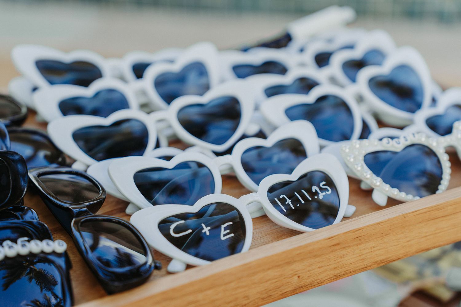 White heart-shaped sunglasses with personalized wedding date and initials arranged on a wooden tray as favors.