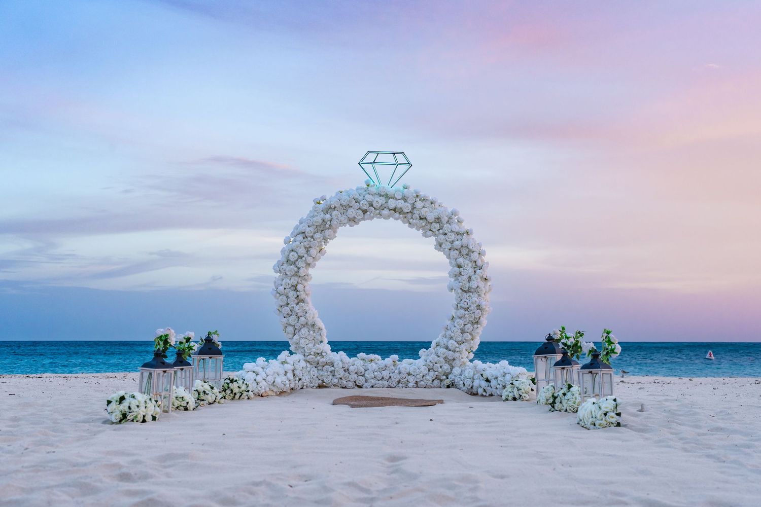 Elegant white floral diamond ring arch installation on a beach at dusk with ocean views in the background.