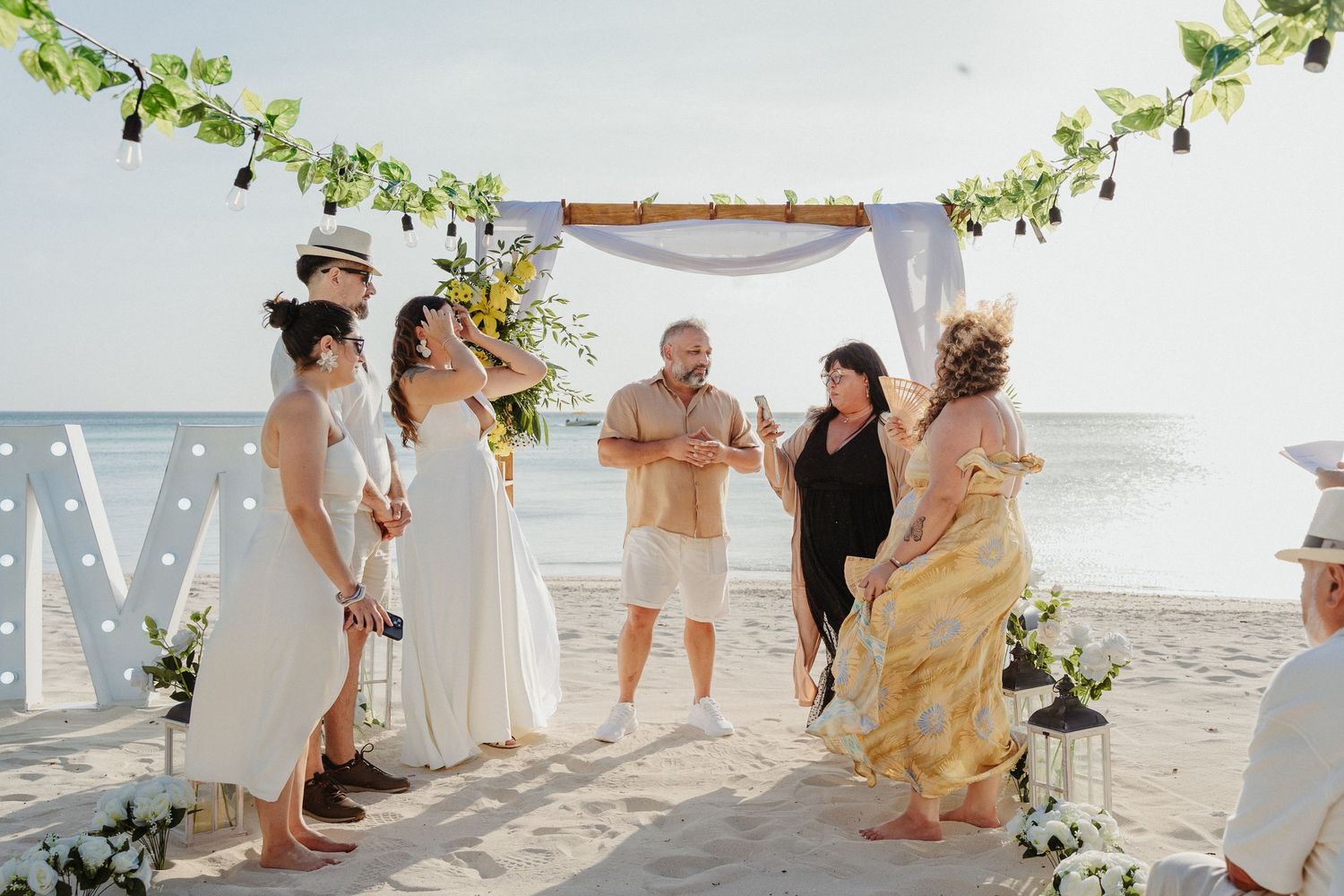 Outdoor beach wedding ceremony with couple and guests under bamboo arch decorated with greenery by the ocean.