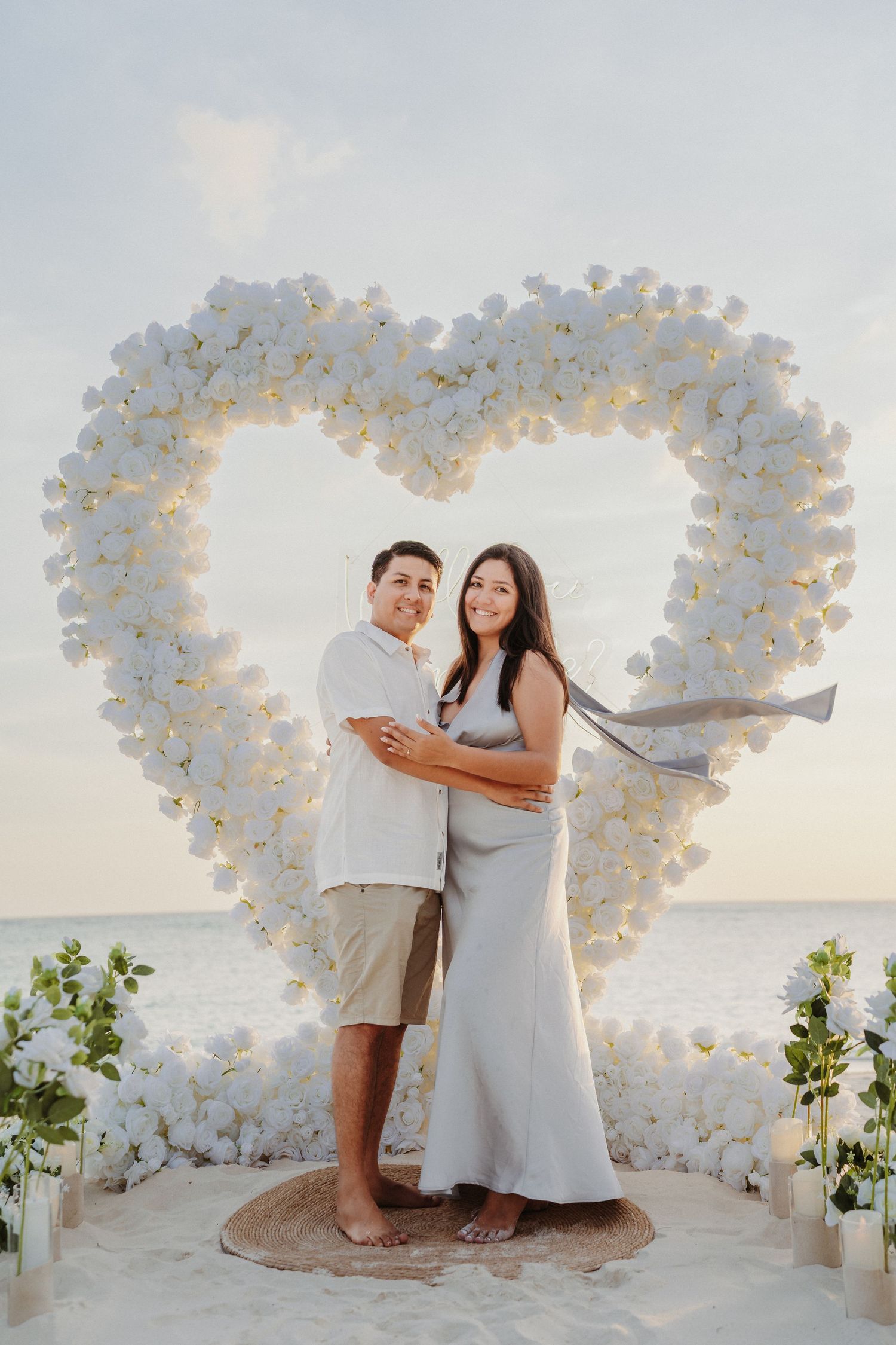 Smiling couple stands before a white rose heart arch on a beach at dusk.