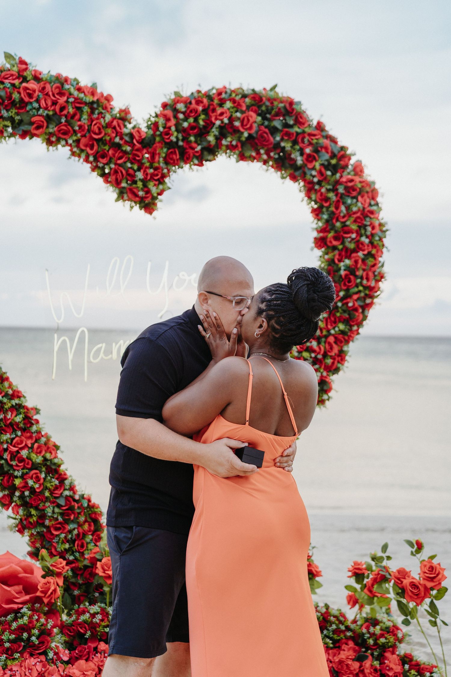 Newly engaged couple shares first kiss under red rose heart arch with Will You Marry Me sign.