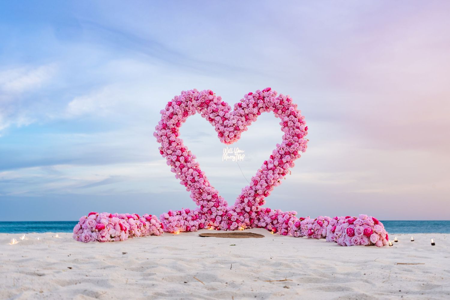 Pink floral heart arch on sandy beach with pastel sky, perfect for romantic wedding ceremony decoration.