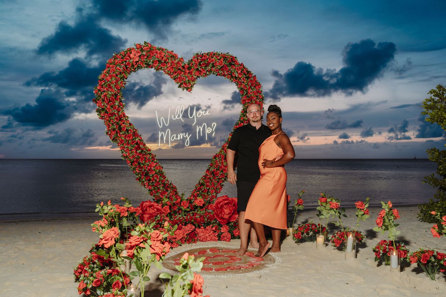 Happy couple poses in front of red rose heart arch with Will You Marry Me sign at sunset.
