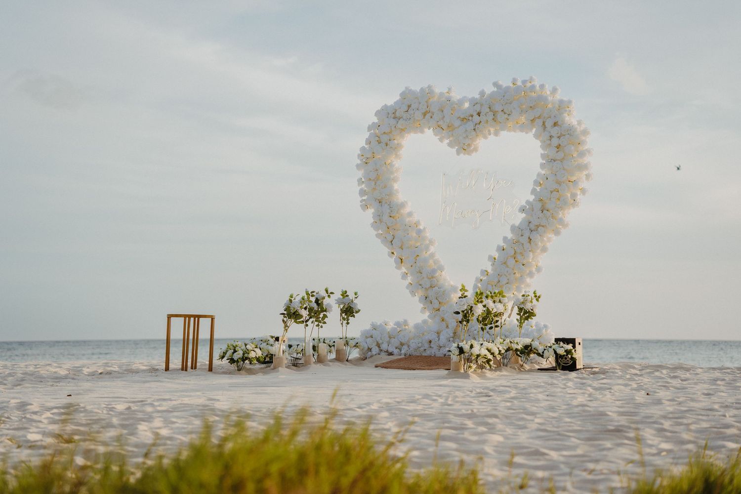 Large white heart-shaped floral arch with flower arrangements on a scenic beach.