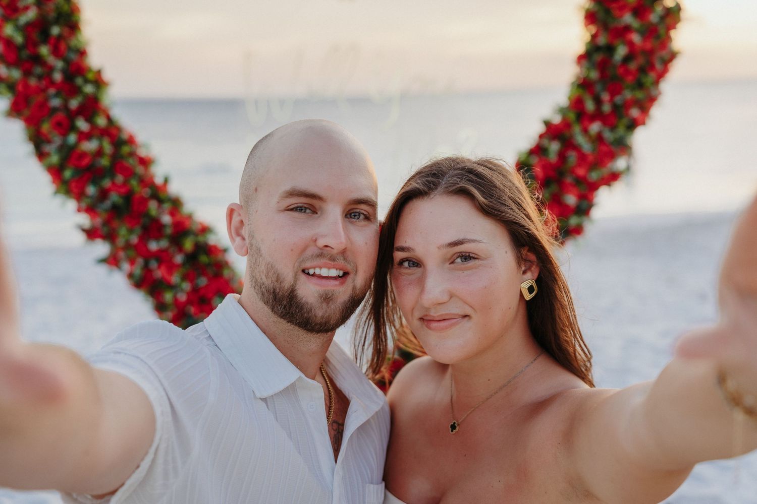 Happy couple takes a selfie in front of a red floral heart arch on beach.