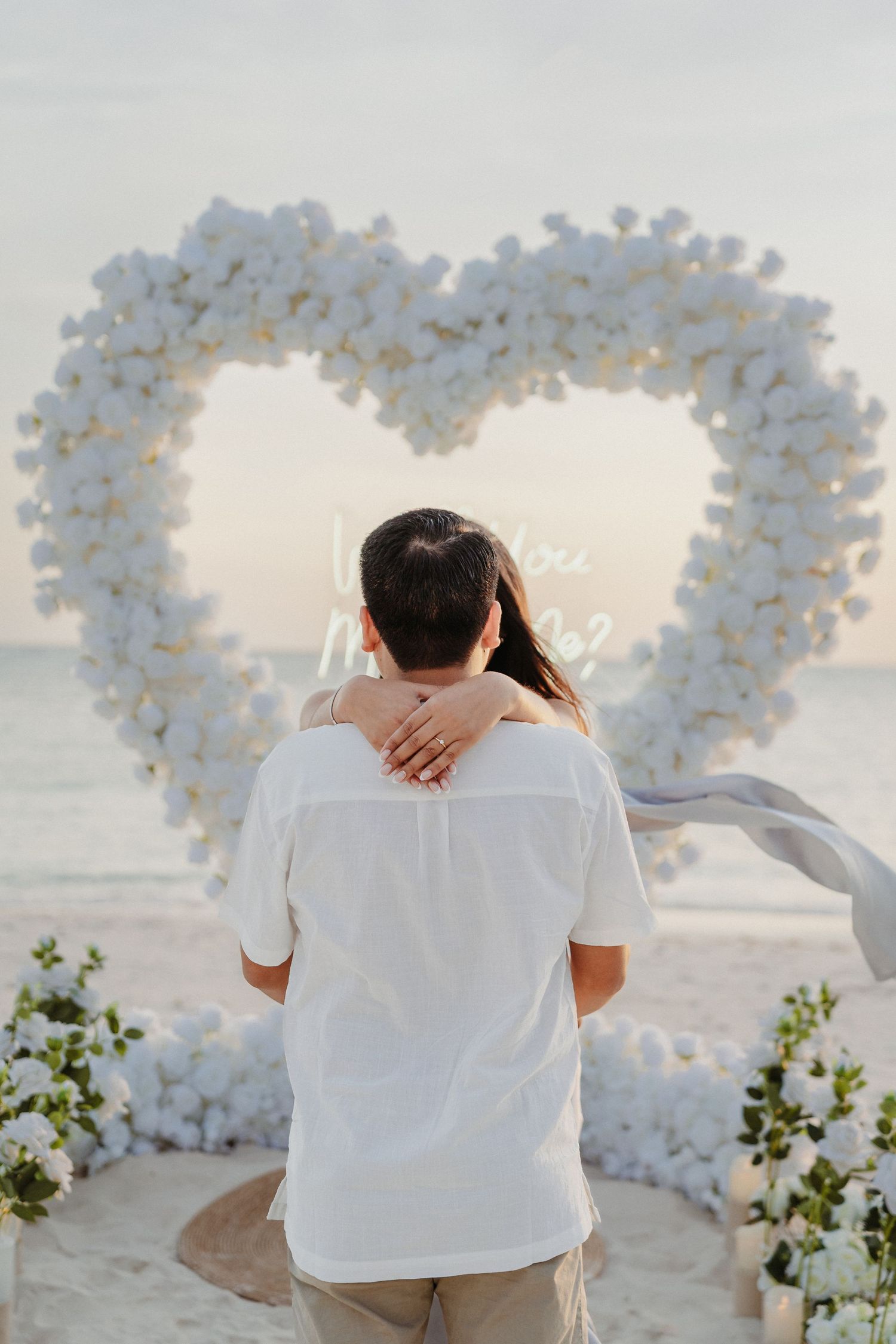 Couple embraces facing a white floral heart arch with neon sign on beach.