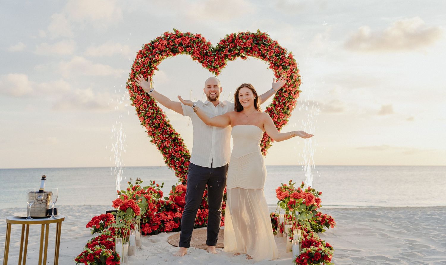 Couple posing joyfully in front of a large red rose heart arch on a beach at sunset.