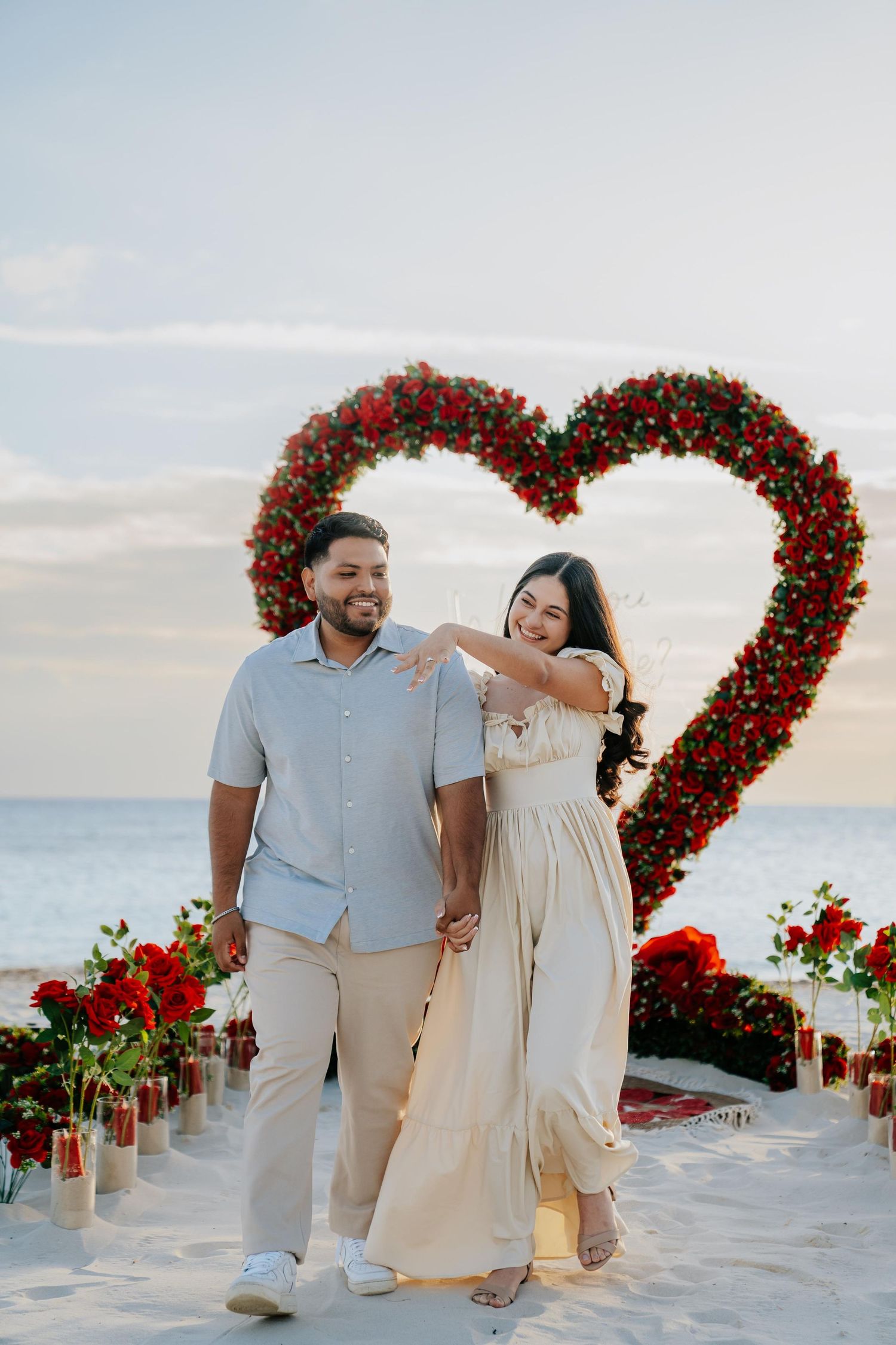Happy couple posing in front of red rose heart-shaped arch at romantic beach proposal setup.