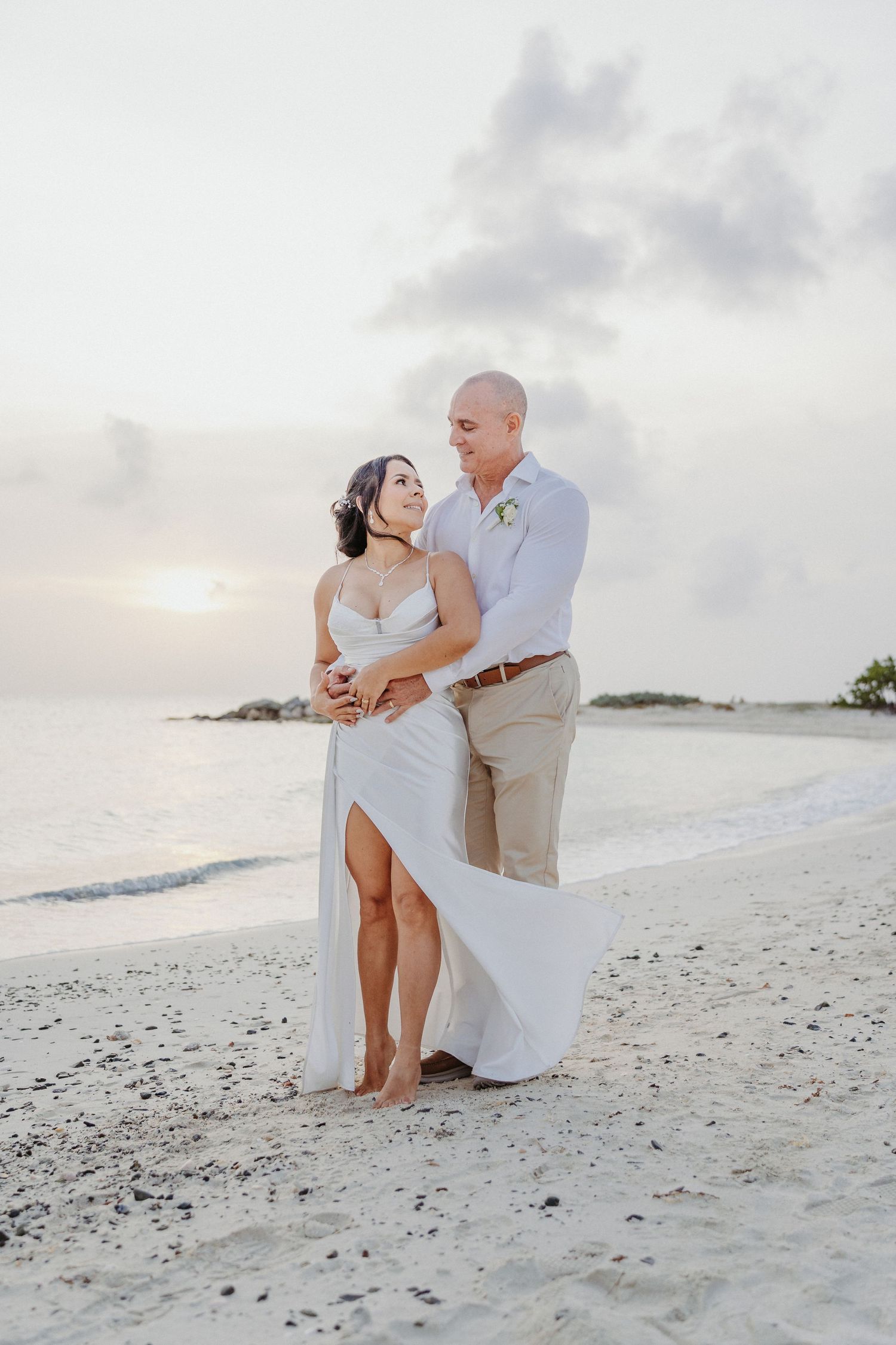 Romantic newlywed couple embracing barefoot on sandy beach at sunset with ocean waves behind them.
