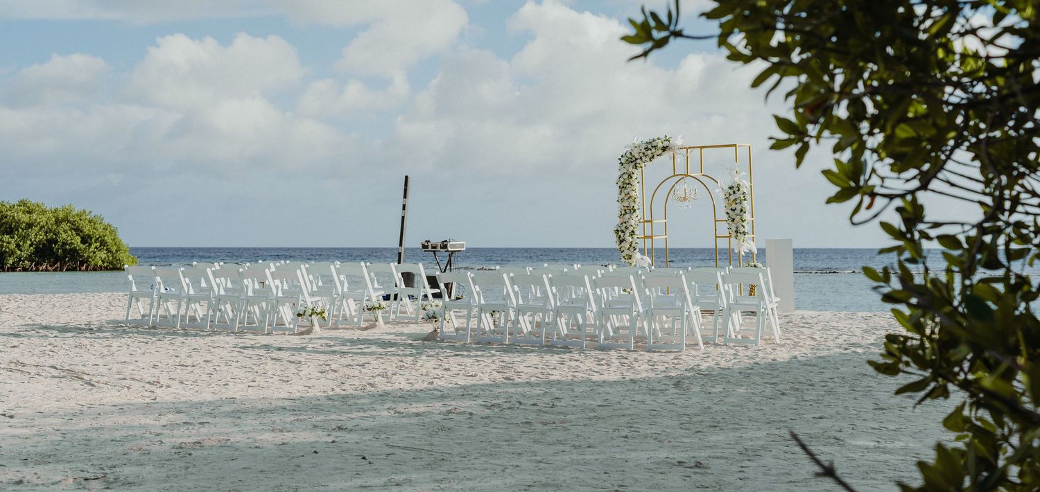 Outdoor beach wedding ceremony setup with white chairs and floral arch by the water.