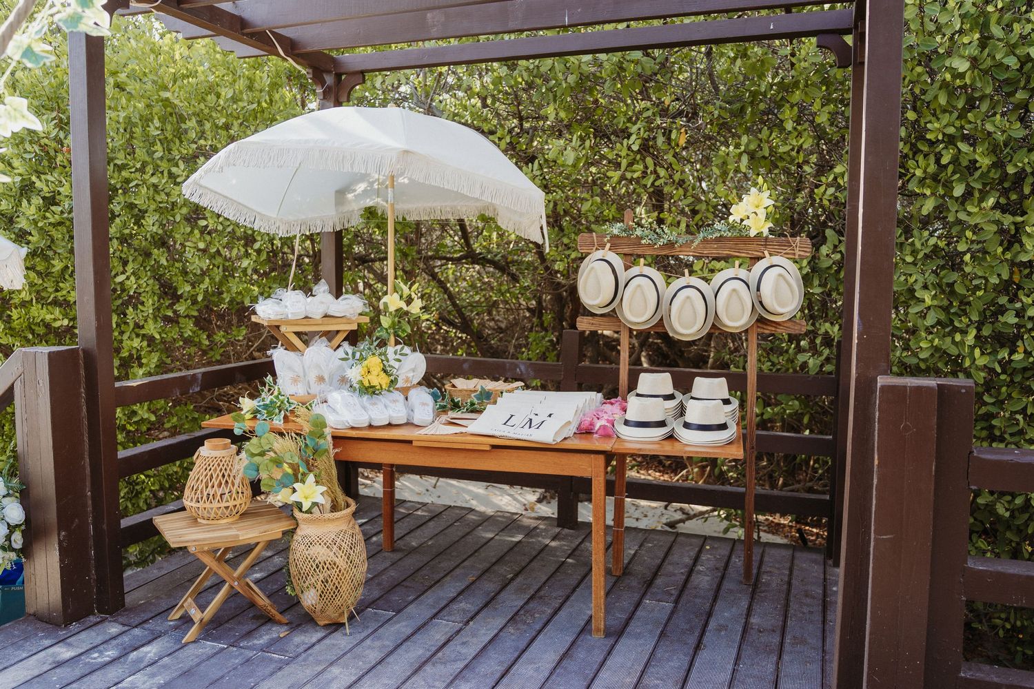 Elegant outdoor garden party setup with white parasol, wooden table, floral arrangements on a deck under a pergola.