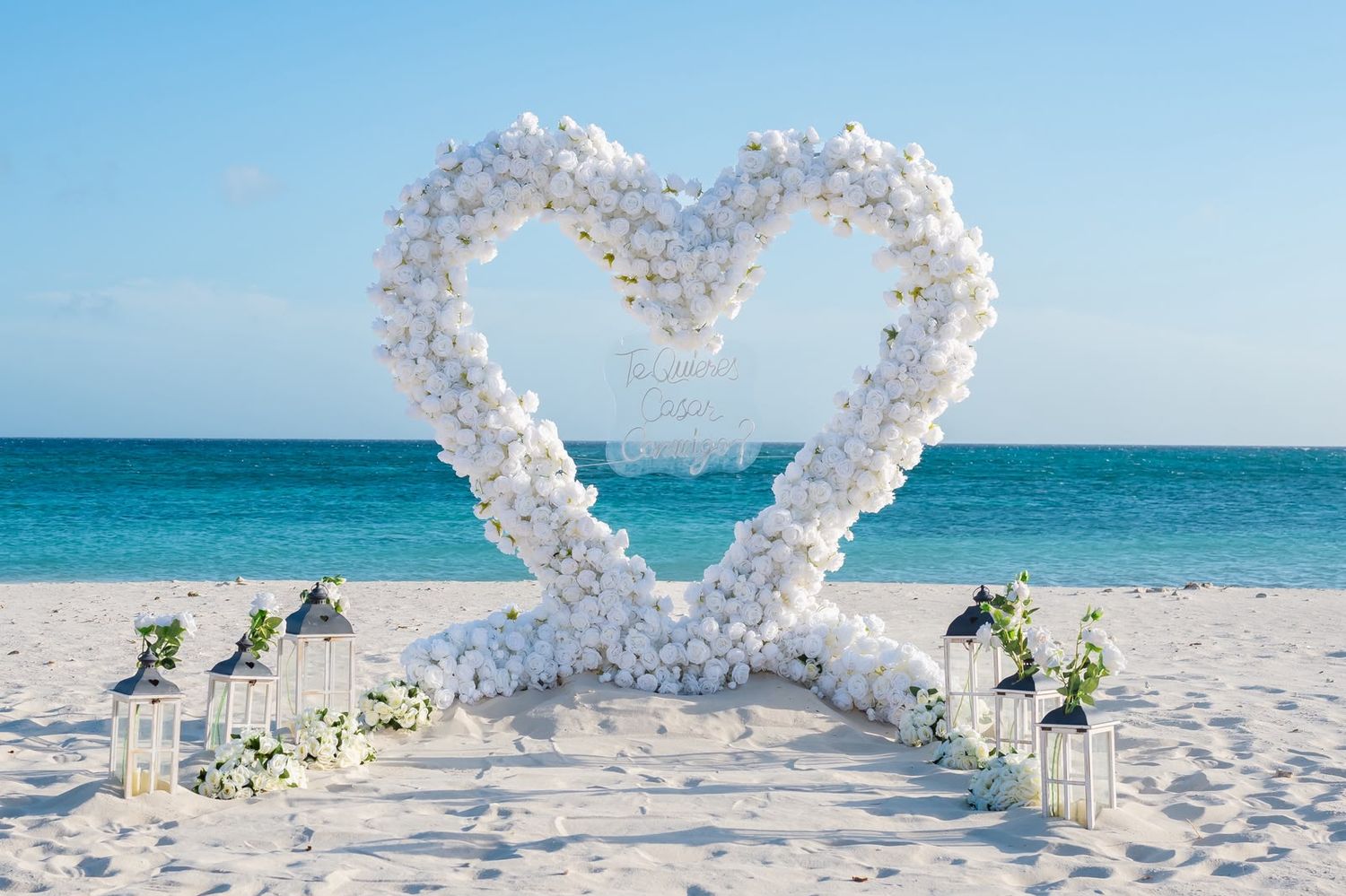 White floral heart-shaped wedding arch with lanterns and flower arrangements on a sandy beach overlooking the turquoise ocean.