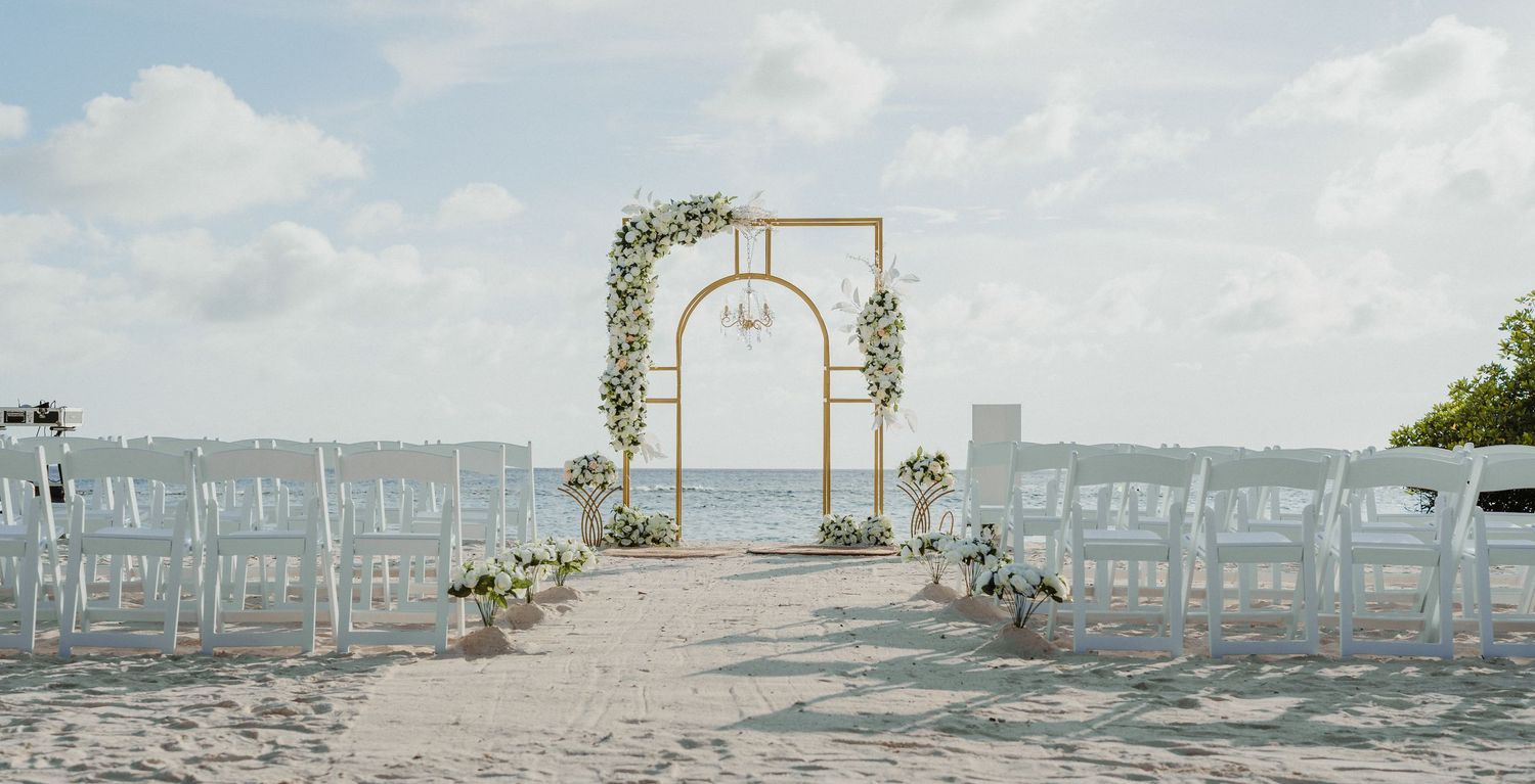 Gold wedding arch with white floral decor and chandelier on a white sandy beach.
