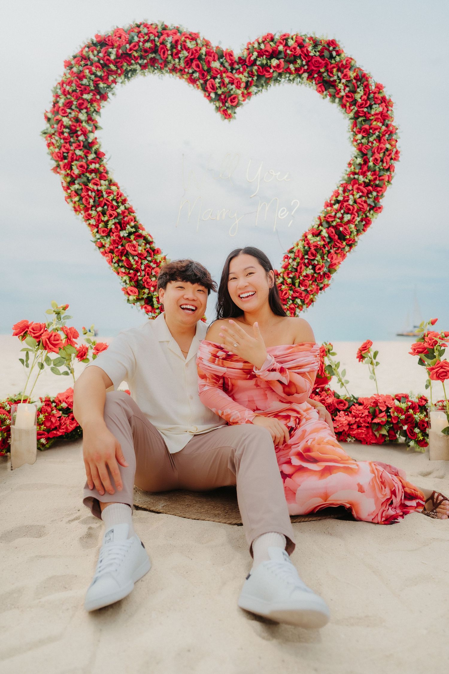 Happy couple laughing in front of large red rose heart arch on beach, woman in pink dress.
