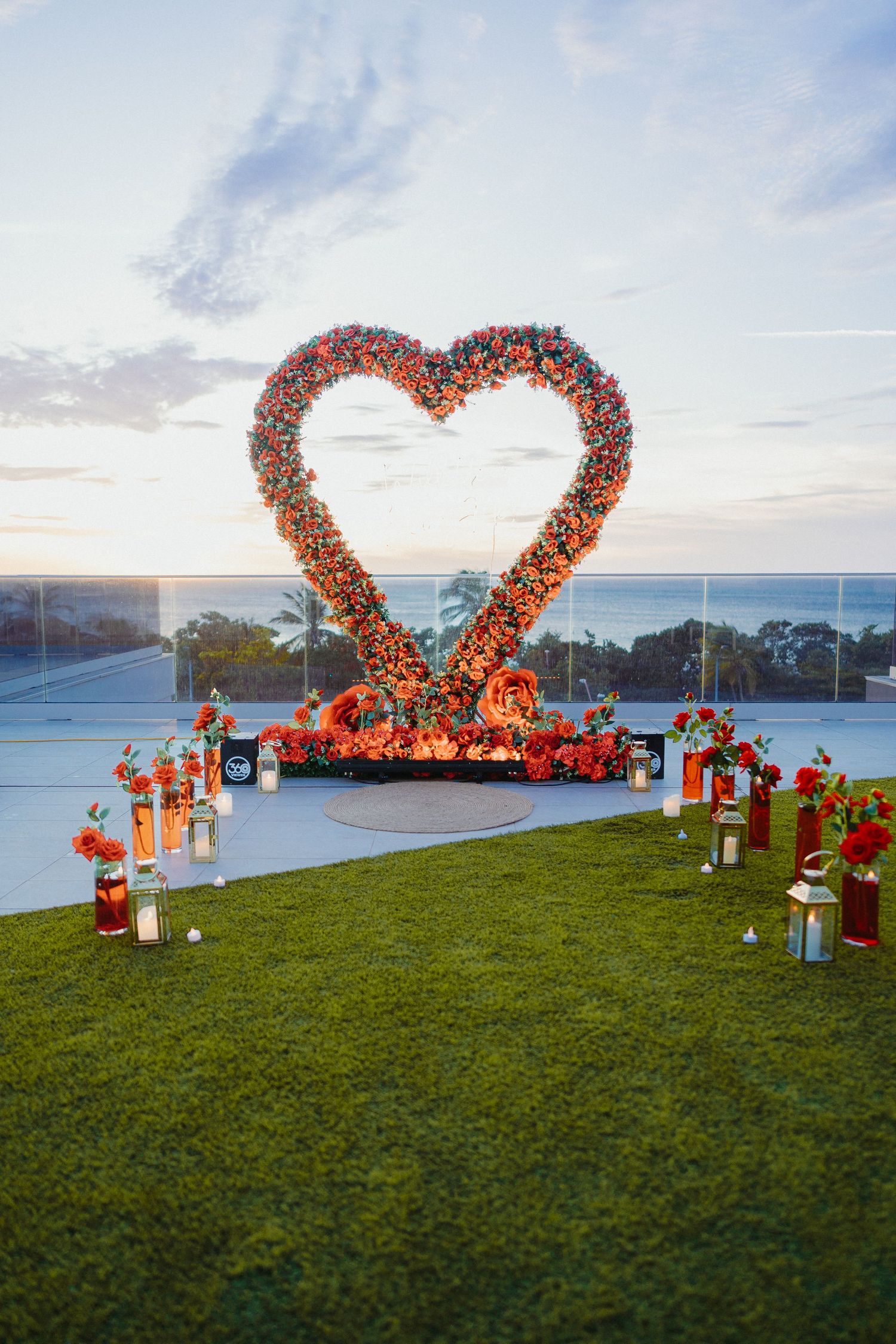 Elegant outdoor marriage proposal setup with large floral heart arch, candles, and flower arrangements at sunset.
