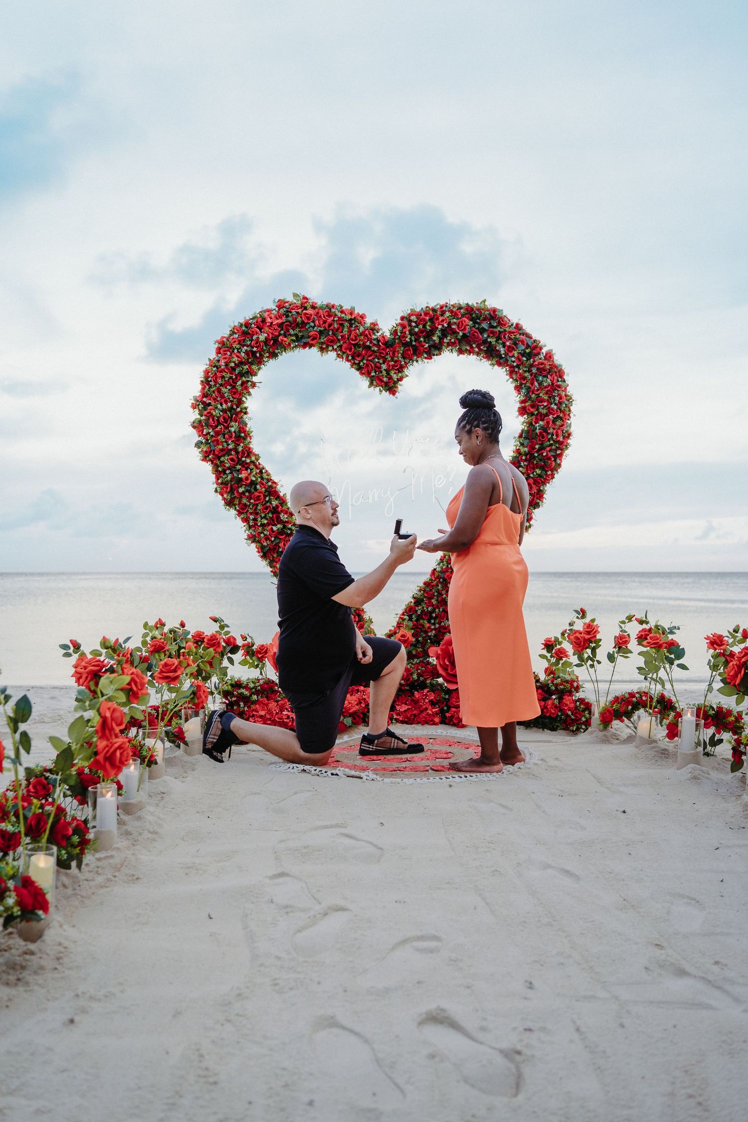 Man kneels to propose to woman in orange dress in front of red rose heart arch on beach.