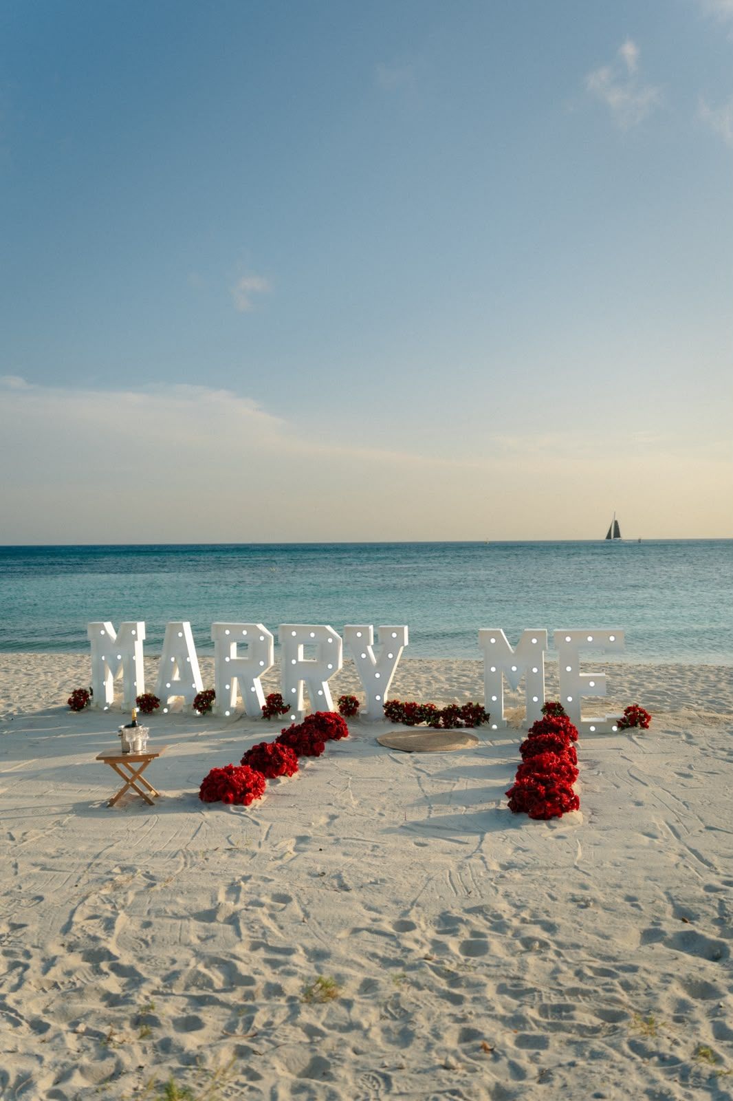 Marry Me marquee letters and red rose petals on a white sand beach at sunset, with a sailboat on the turquoise ocean.