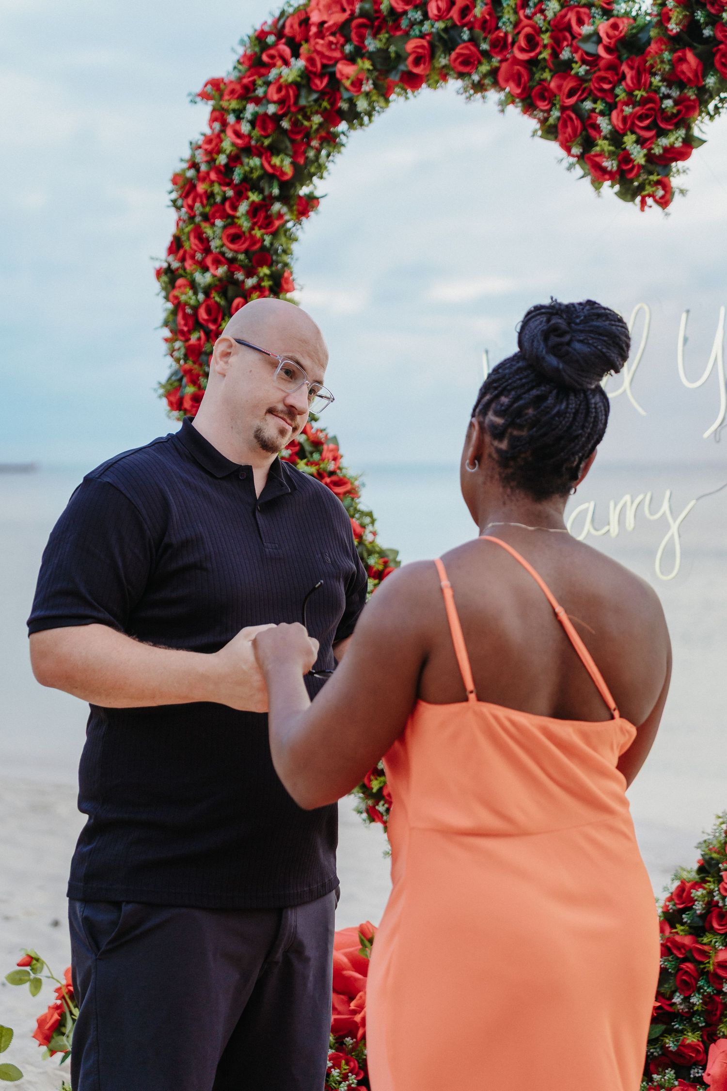 Man in black shirt holds ring box while proposing to woman in orange dress on beach.