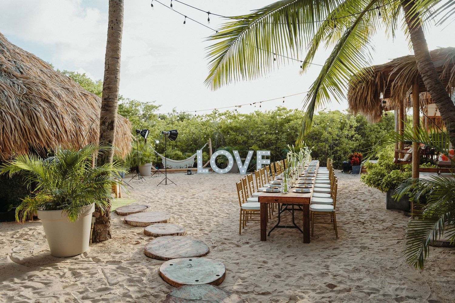 Tropical beach wedding reception setup with long dining table, string lights, and large LOVE letter sign.