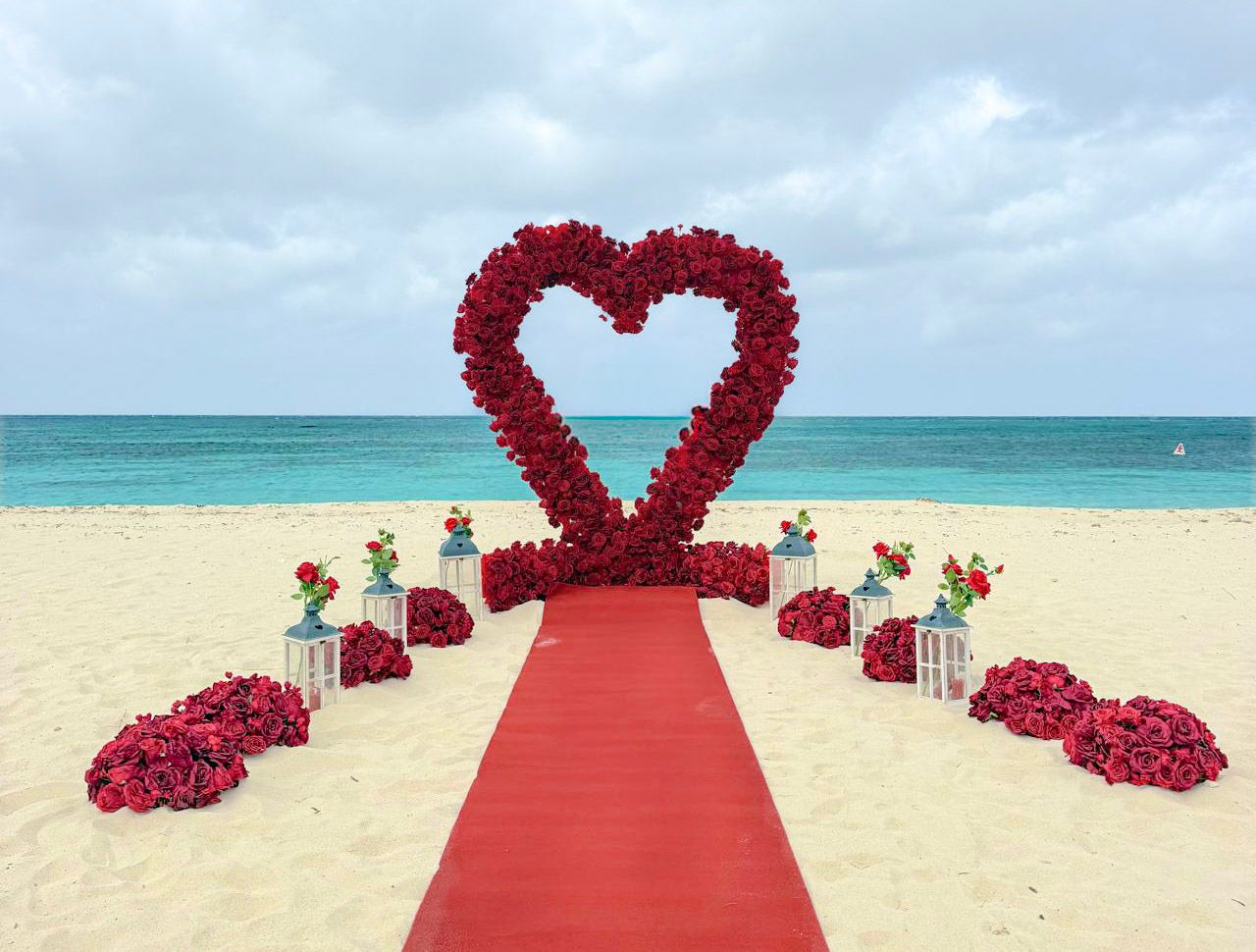 Heart-shaped red rose arch and red carpet aisle on a sandy tropical beach wedding setup with turquoise ocean and rose petal arrangements.