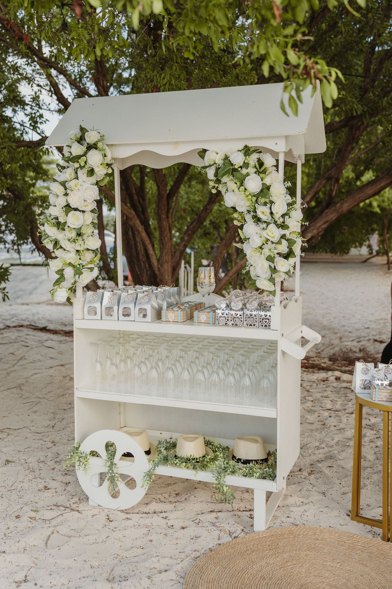 White floral wedding bar cart with wine glasses and white roses, set outdoors on sandy beach under green trees.