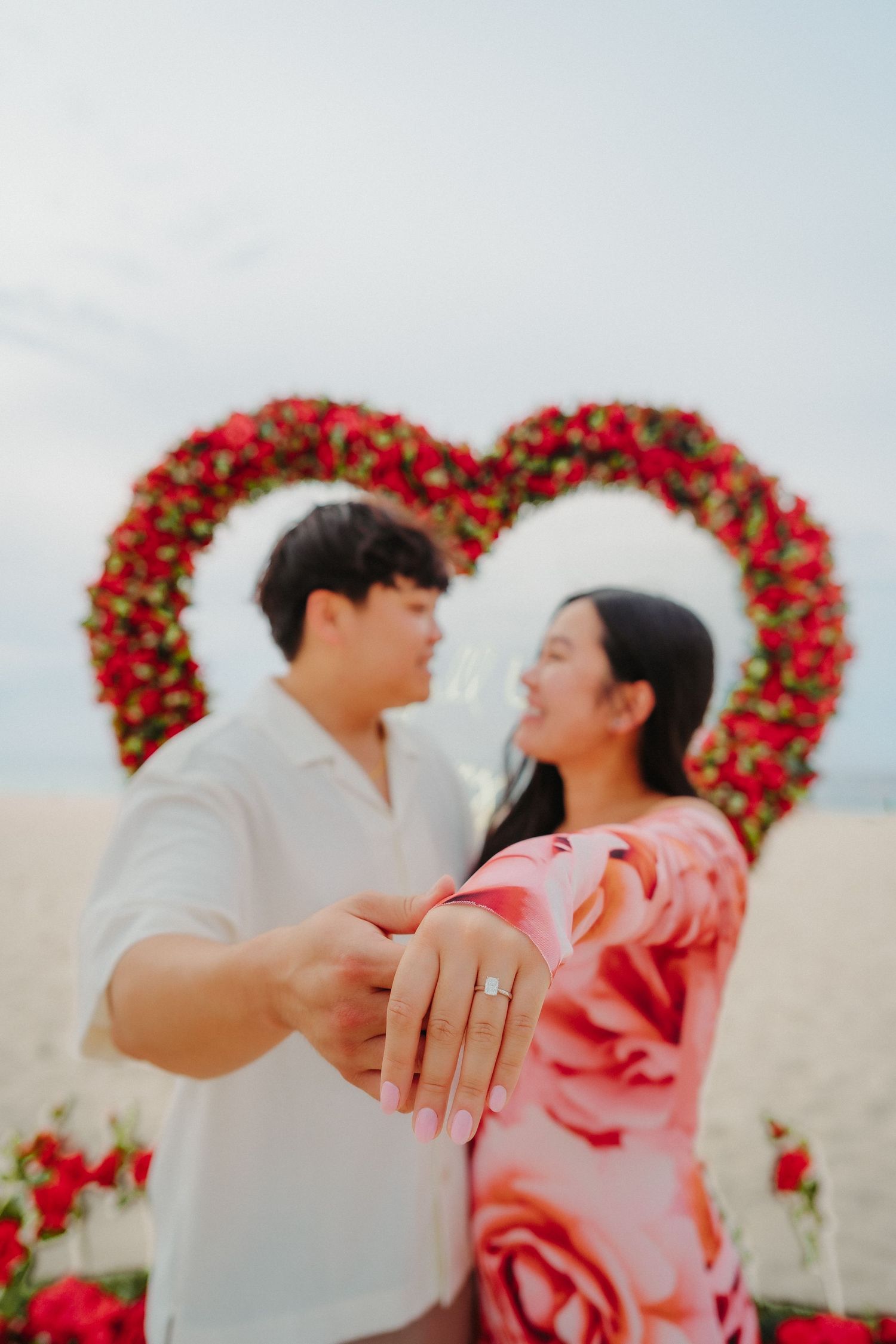 Newly engaged couple showing off engagement ring in front of red rose heart arch on beach.