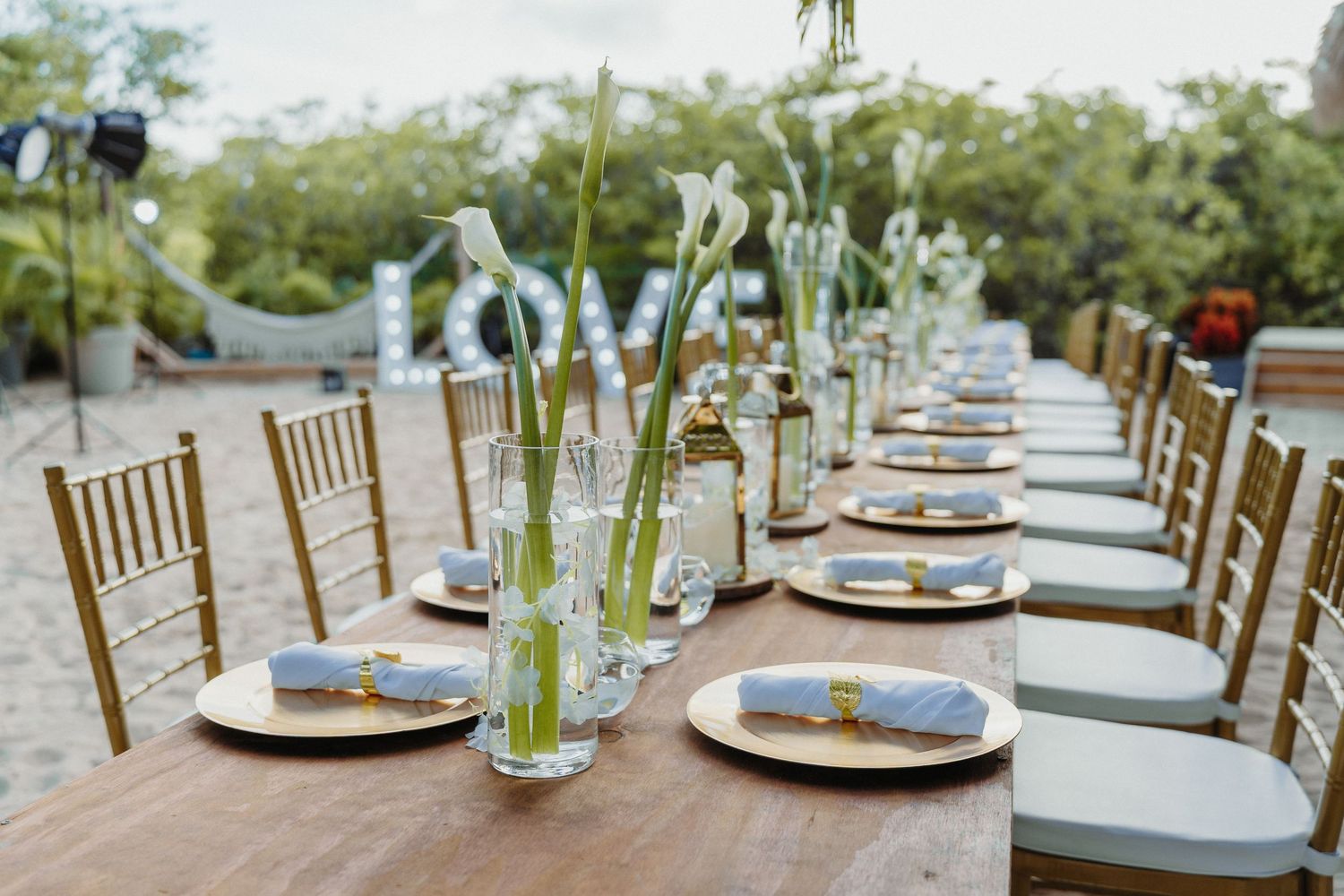 Rustic outdoor wedding reception table with tall calla lily centerpieces, gold chargers, and LOVE marquee sign.