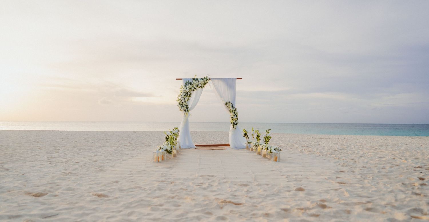 White floral wedding arch set on pristine sandy beach at soft pastel sunset.