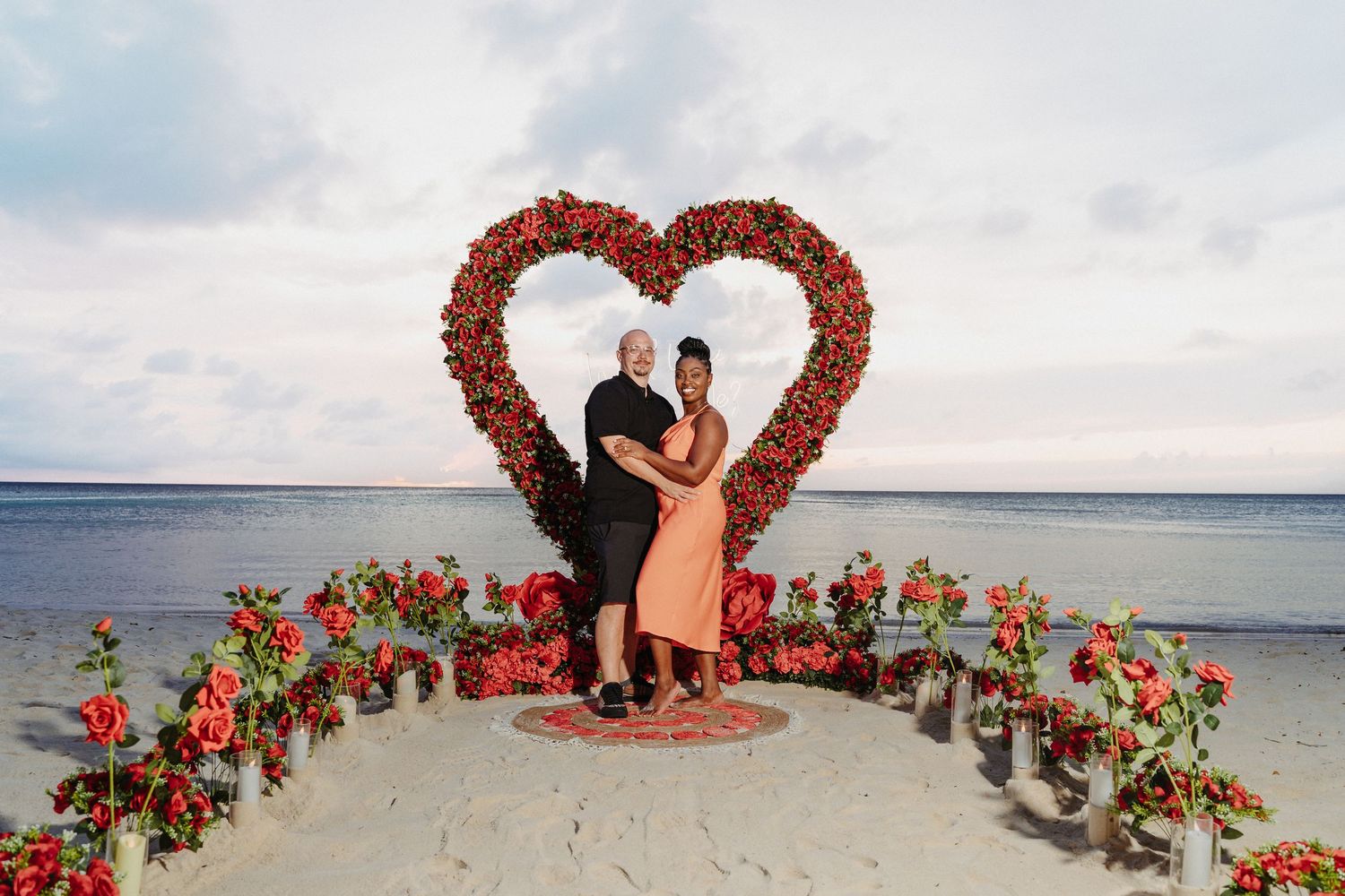Couple poses in front of a red rose heart arch on a beach at sunset.