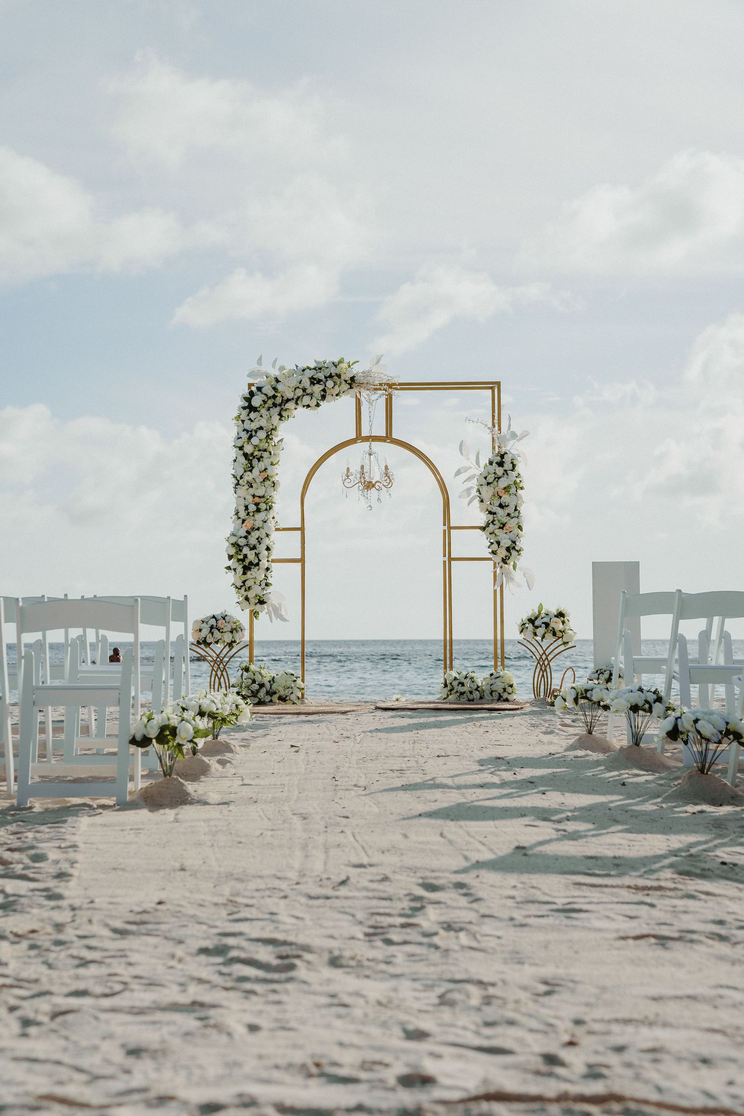 Elegant gold wedding arch with white flowers on a sandy beach overlooking the ocean.